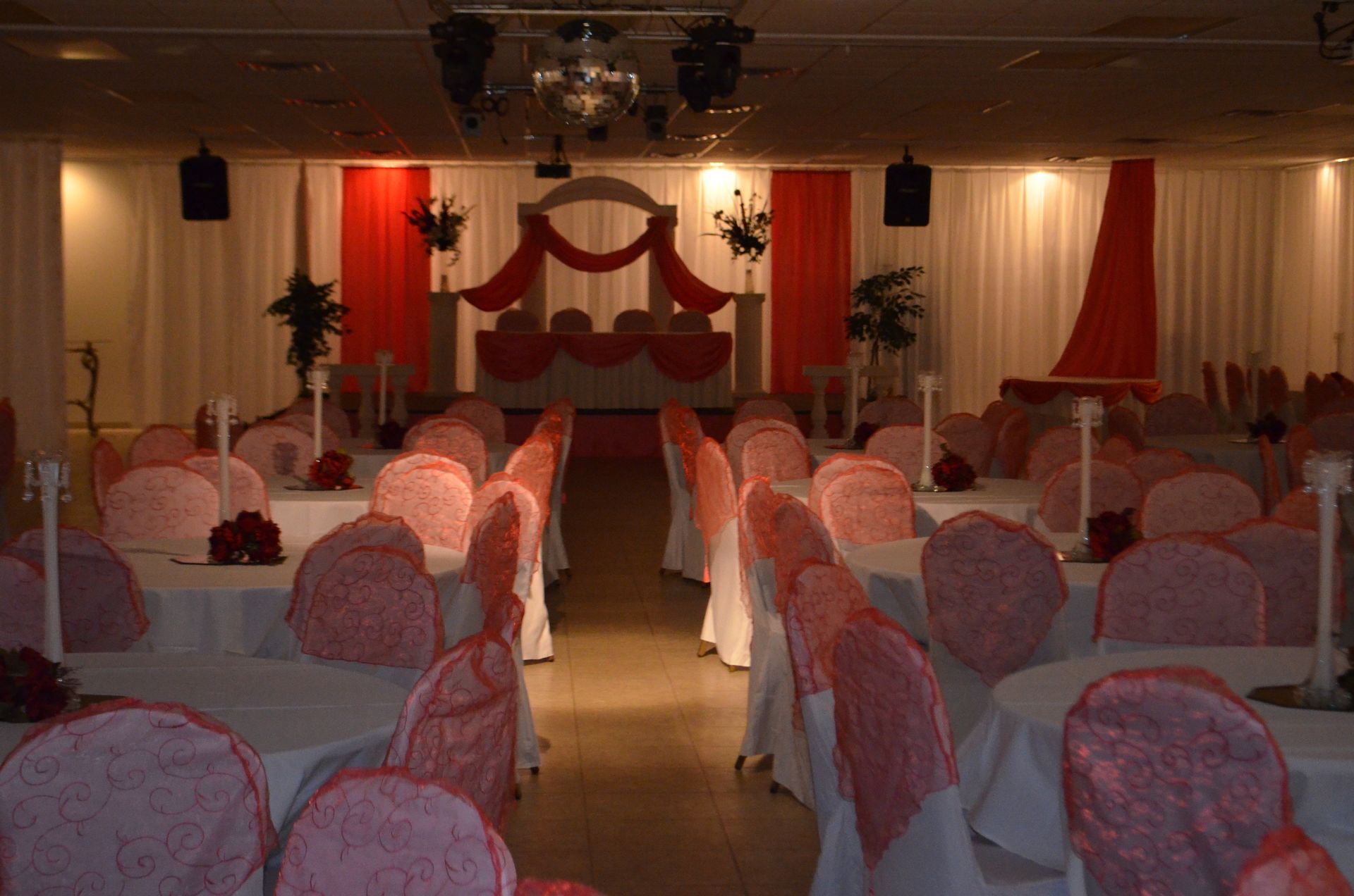 Wedding reception hall with pink-covered chairs facing a decorated stage and red drapes