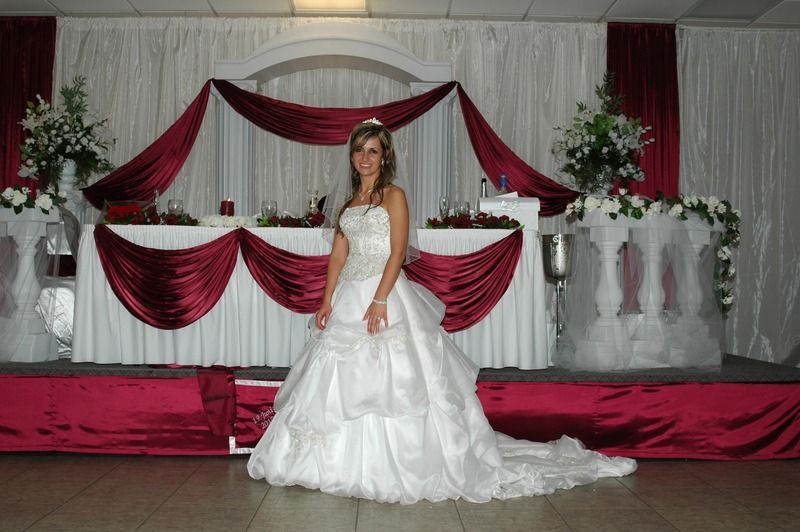 Woman in a white gown standing on a decorated stage with red drapes and floral arrangements