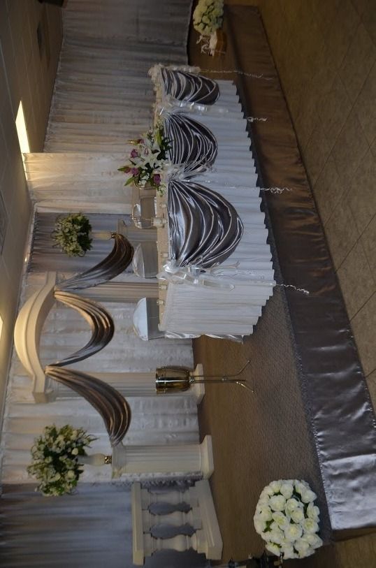 Decorated stage with white draping, floral arrangements, silver arches, and a covered central table at an event.
