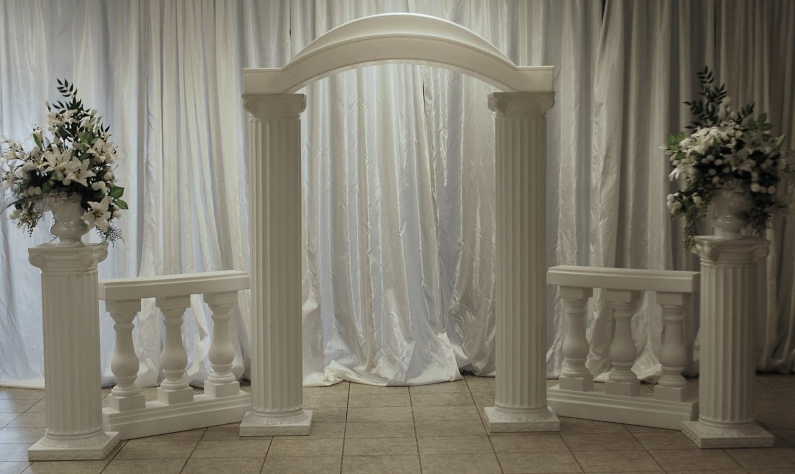 Elegant white wedding arch with draped curtains and floral arrangements on pedestals