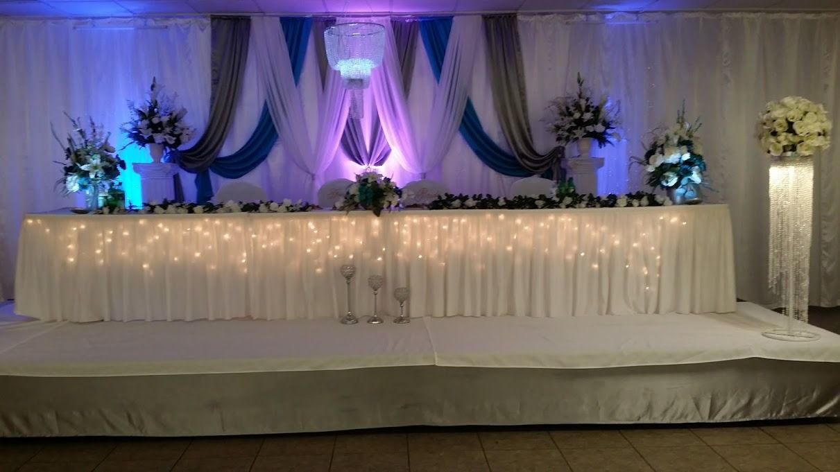 Decorated wedding stage with white drapes, purple lighting, floral arrangements, and a long head table