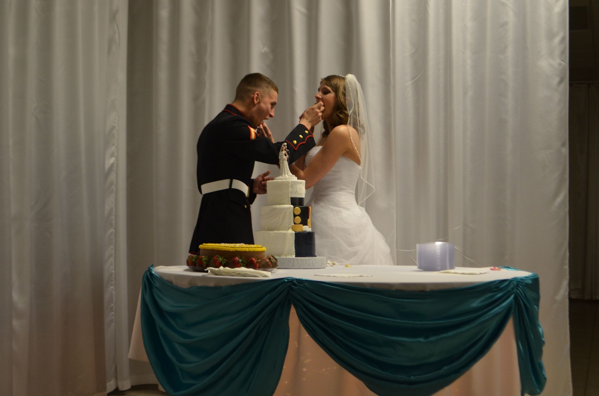 Bride and groom share cake at a wedding reception table with teal draping