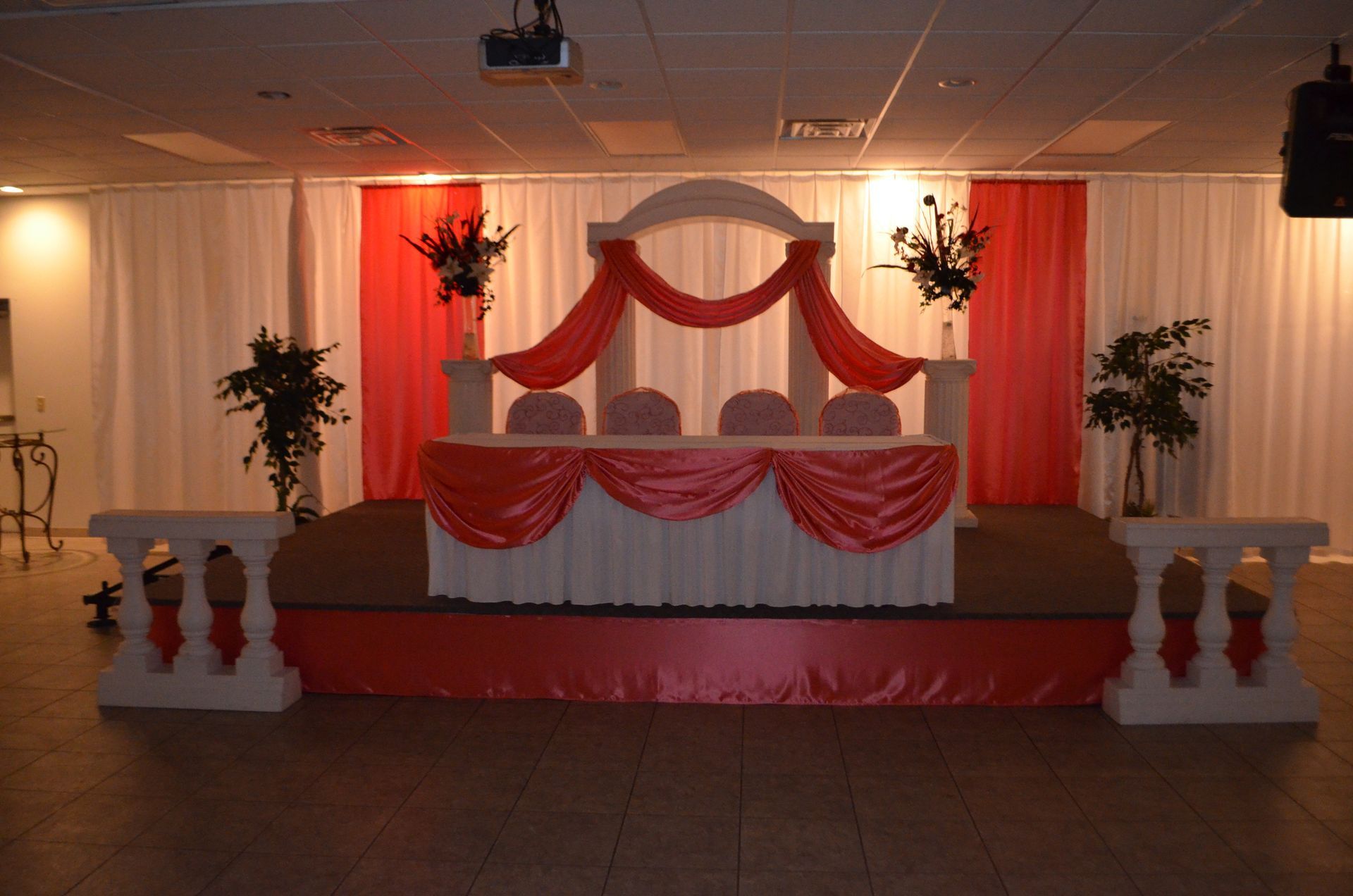 Decorated wedding stage with red-and-white drapes, floral arrangements, and chairs in a banquet hall