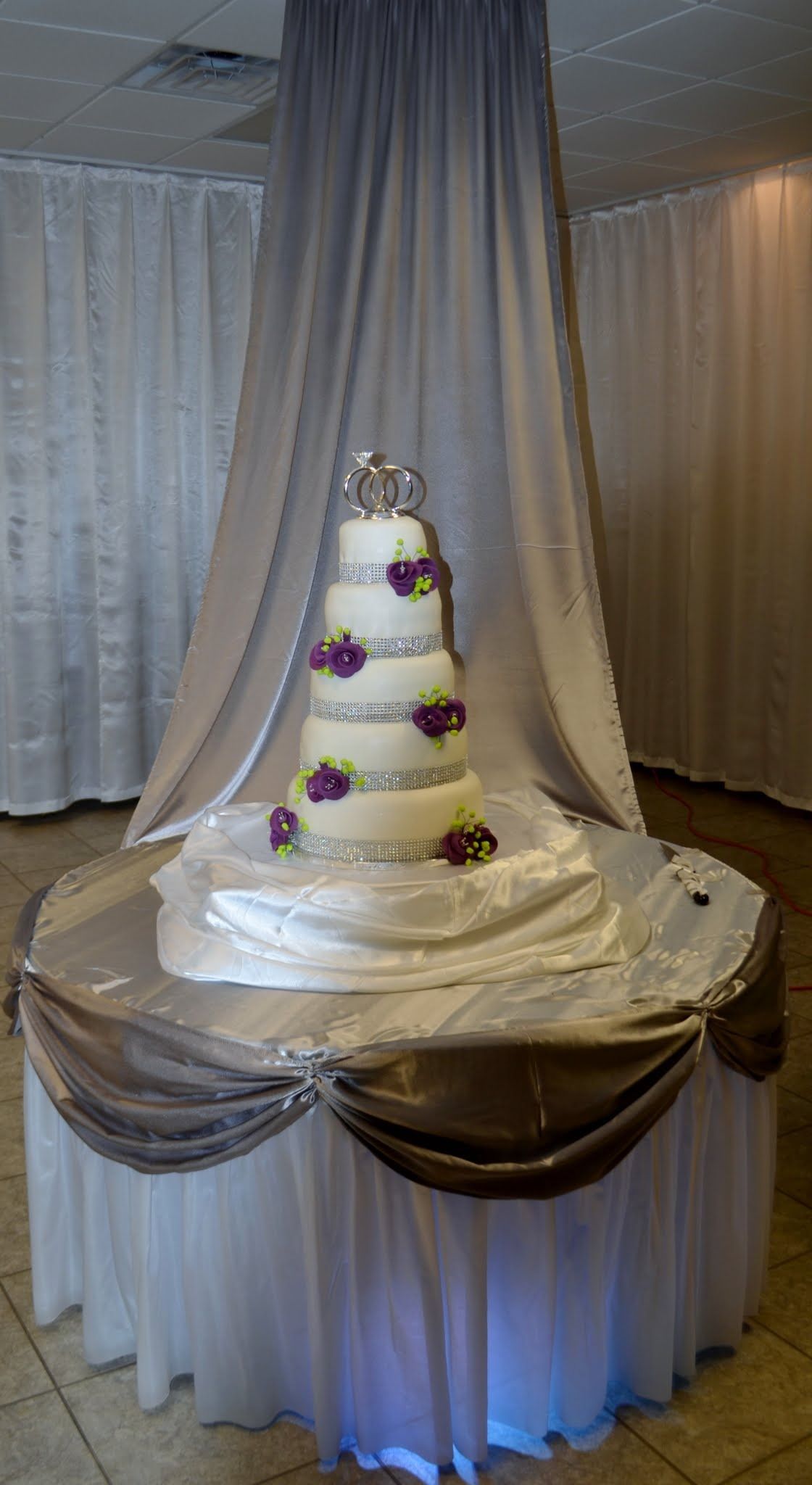 Tiered white wedding cake with purple flowers on a draped round table in a decorated reception hall