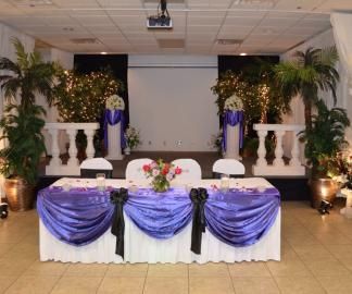 Decorated banquet table with purple drapes and floral centerpiece on a stage with plants and white columns