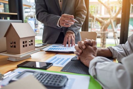 Real estate meeting with a house model, documents, calculator, and hands discussing a contract on a table Real estate meeting with a house model, documents, calculator, and hands discussing a contract on a table