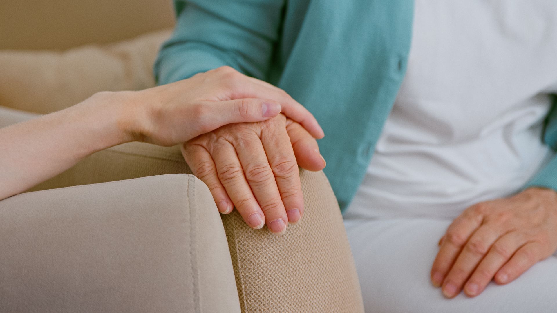 A hand resting gently on another person’s hand on a beige armchair, suggesting comfort and support.
