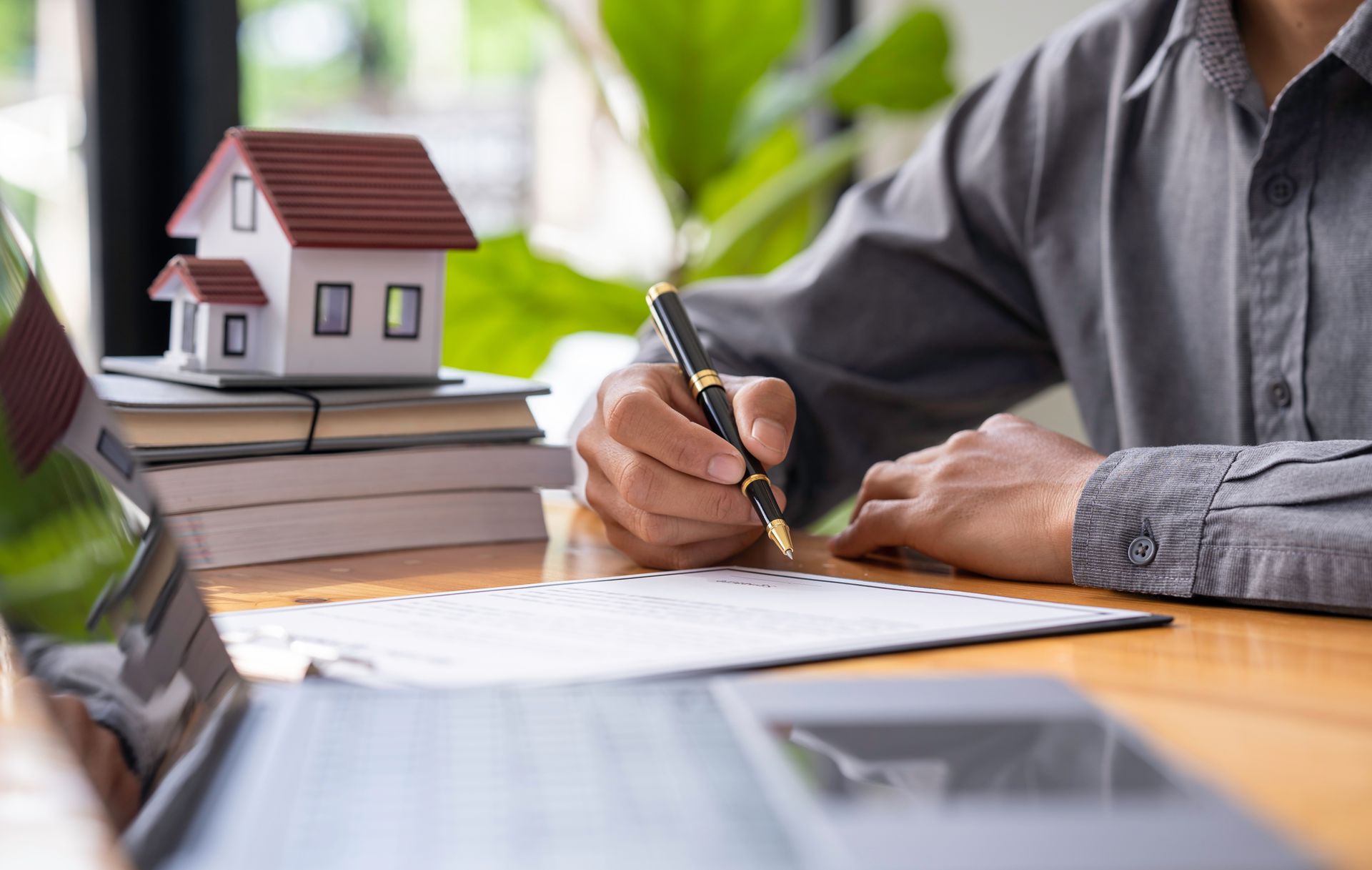Person writing at a desk beside a small model house and stacked books in a bright office setting Person writing at a desk beside a small model house and stacked books in a bright office setting