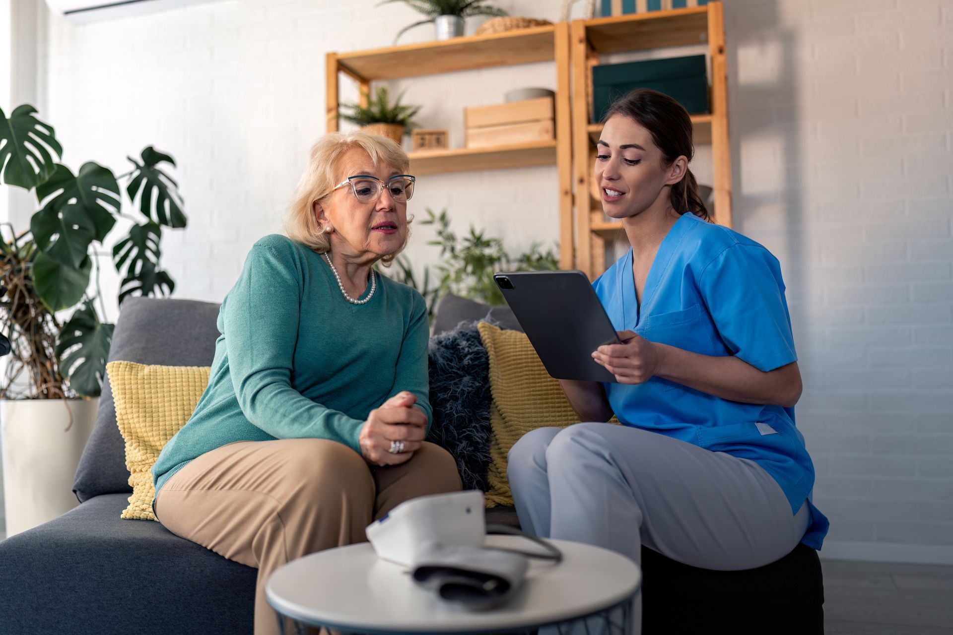 Nurse reviewing a tablet with a seated patient in a bright living room, with a blood pressure monitor on the table