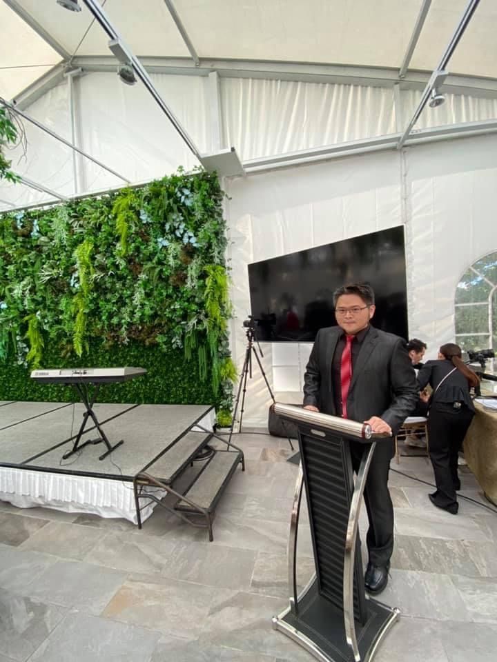 A man in a suit and red tie is standing at a podium in front of a green wall.