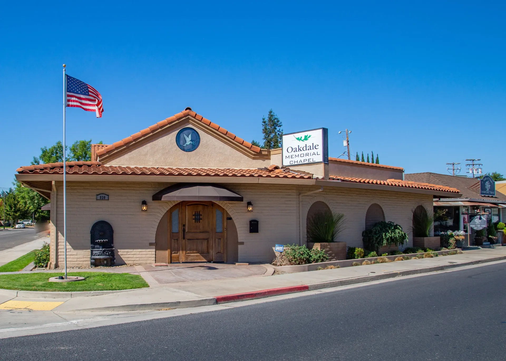 Adobe-style building with American flag, arched wooden entrance, and white sign under a clear blue sky