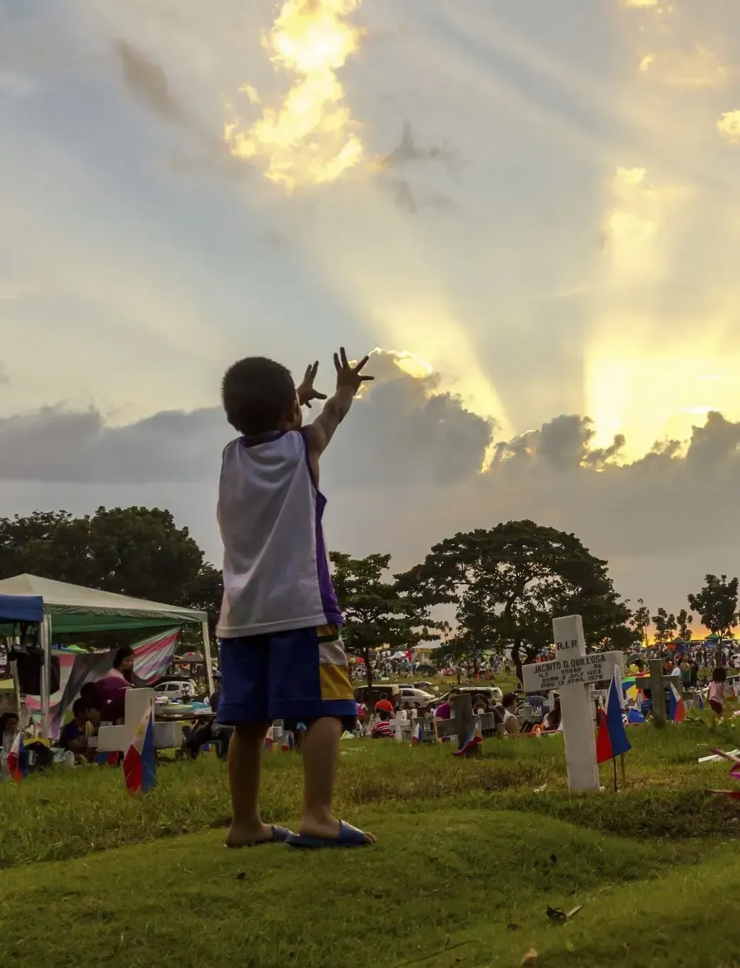 Child on grassy field reaching toward the sky at sunset, with a crowd and tents in the background.