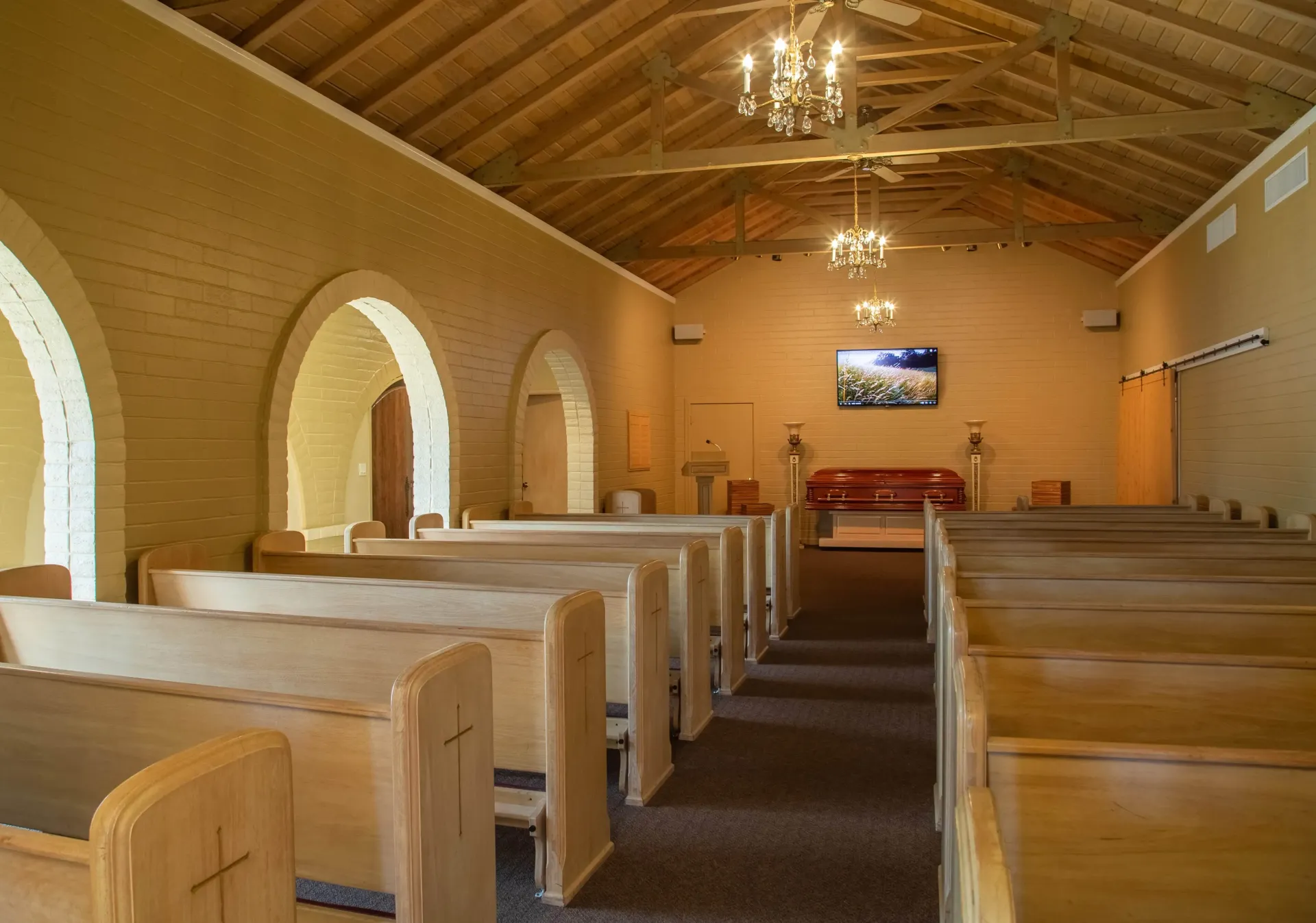 Inside a small church with wooden pews, arched windows, and a chandelier-lit vaulted ceiling