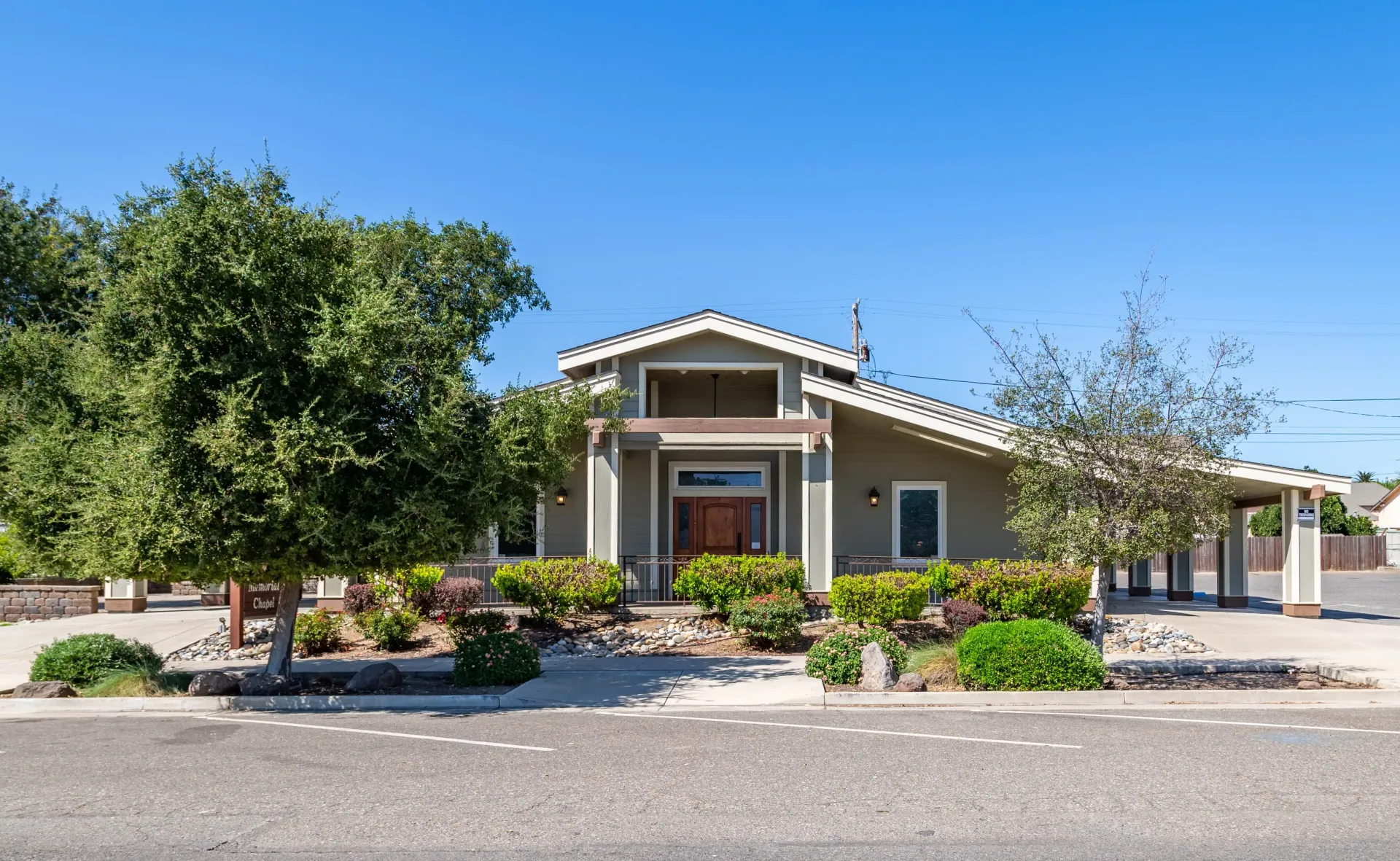 Single-story beige house with a front porch, desert landscaping, and a large tree under a clear blue sky