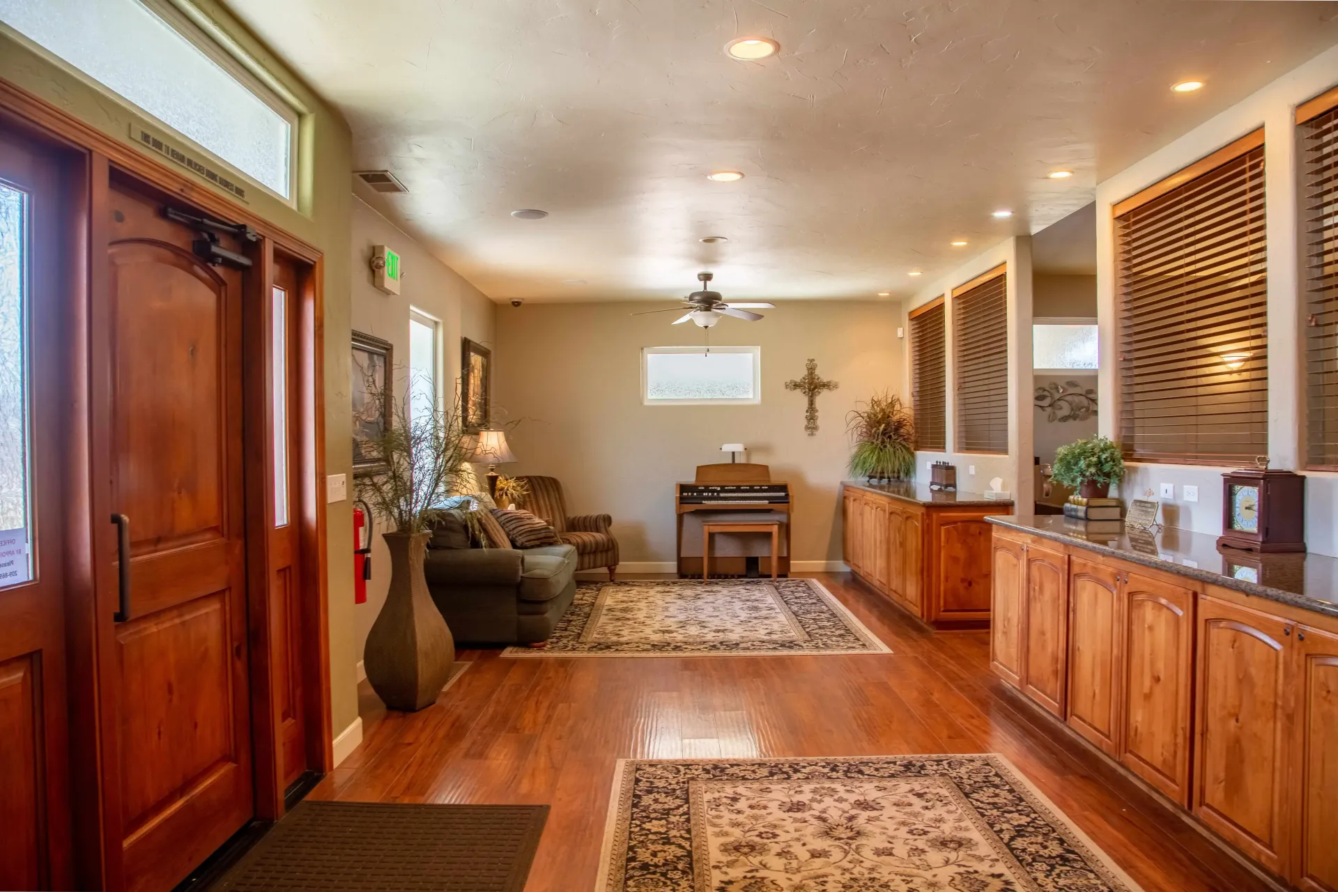 Long hallway with wood cabinets, plants, rugs, piano, and cross in a warmly lit interior.