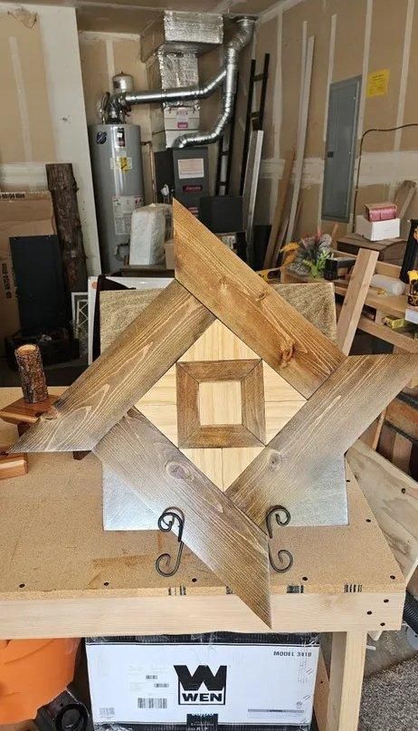 Wooden geometric sculpture on a workbench in a cluttered workshop, with metal hooks below.