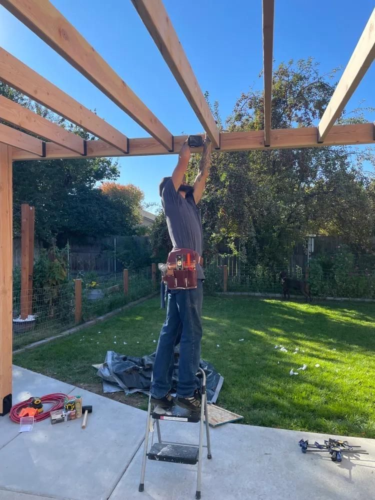 Person on a ladder installing wooden beams on a backyard patio pergola.