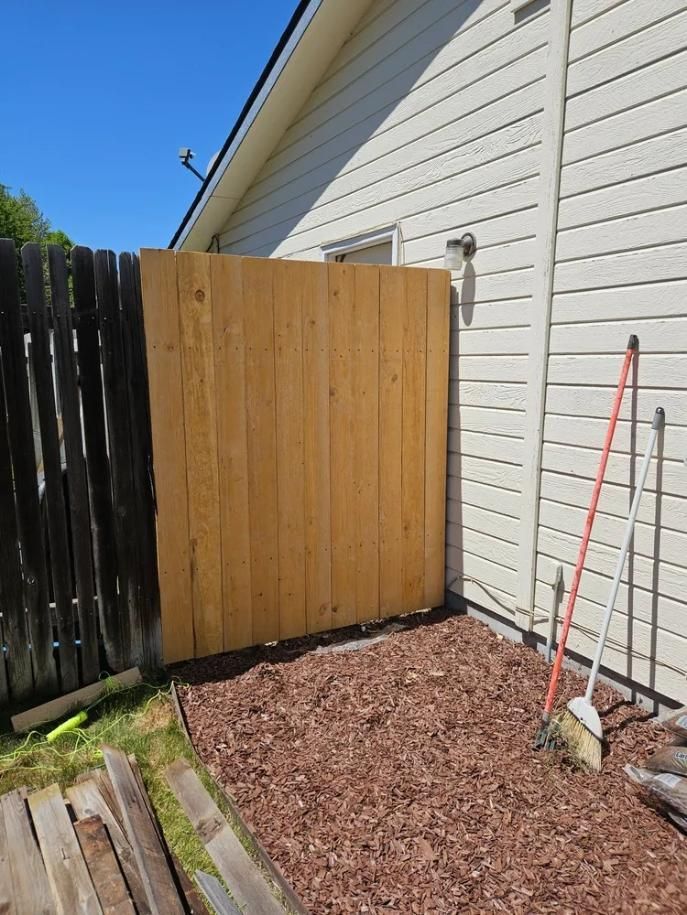 Wooden privacy fence panel beside a house, with a rake leaning against the wall and gravel on the ground.
