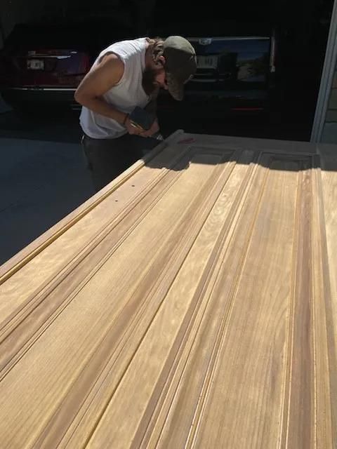 Person inspecting a large wooden surface in a garage or workshop, with parked vehicles in the background