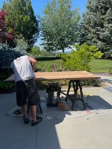 Person sanding a wooden board on sawhorses in a sunny driveway with trees and lawn behind.