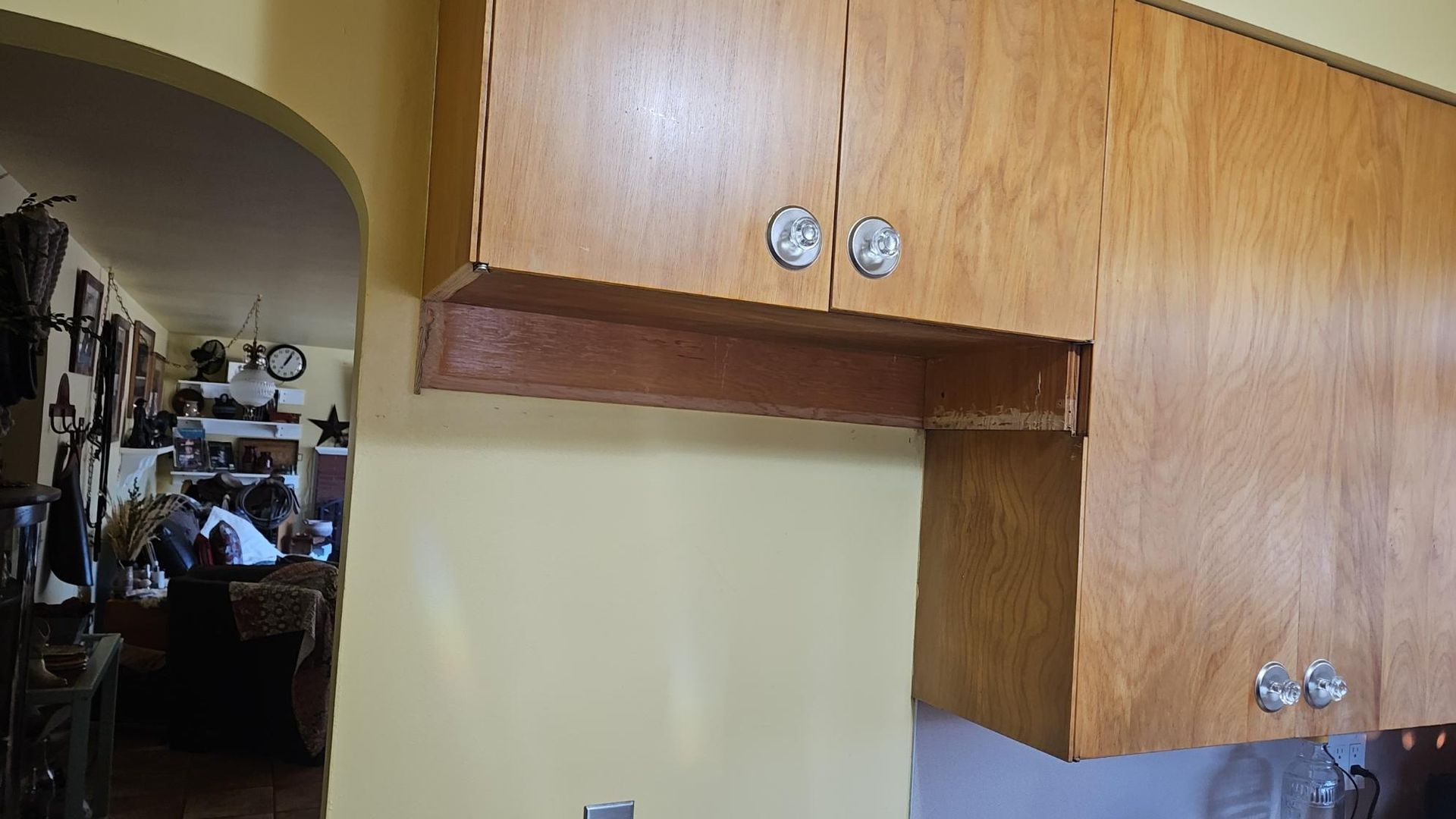 Wooden kitchen cabinets above a cream wall, with an arched doorway leading to a cluttered room.
