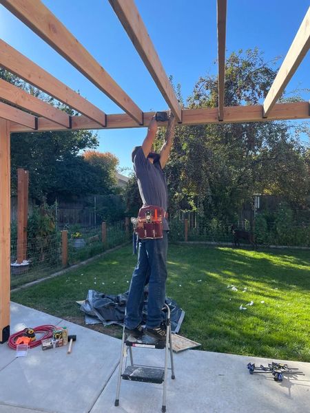 Person standing on a ladder installing wooden beams in a backyard patio pergola construction.