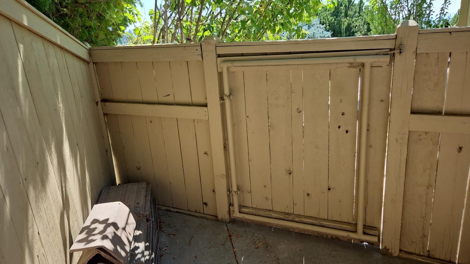 Small fenced patio with tan wooden panels, a covered bench, and sunlight through green trees above.