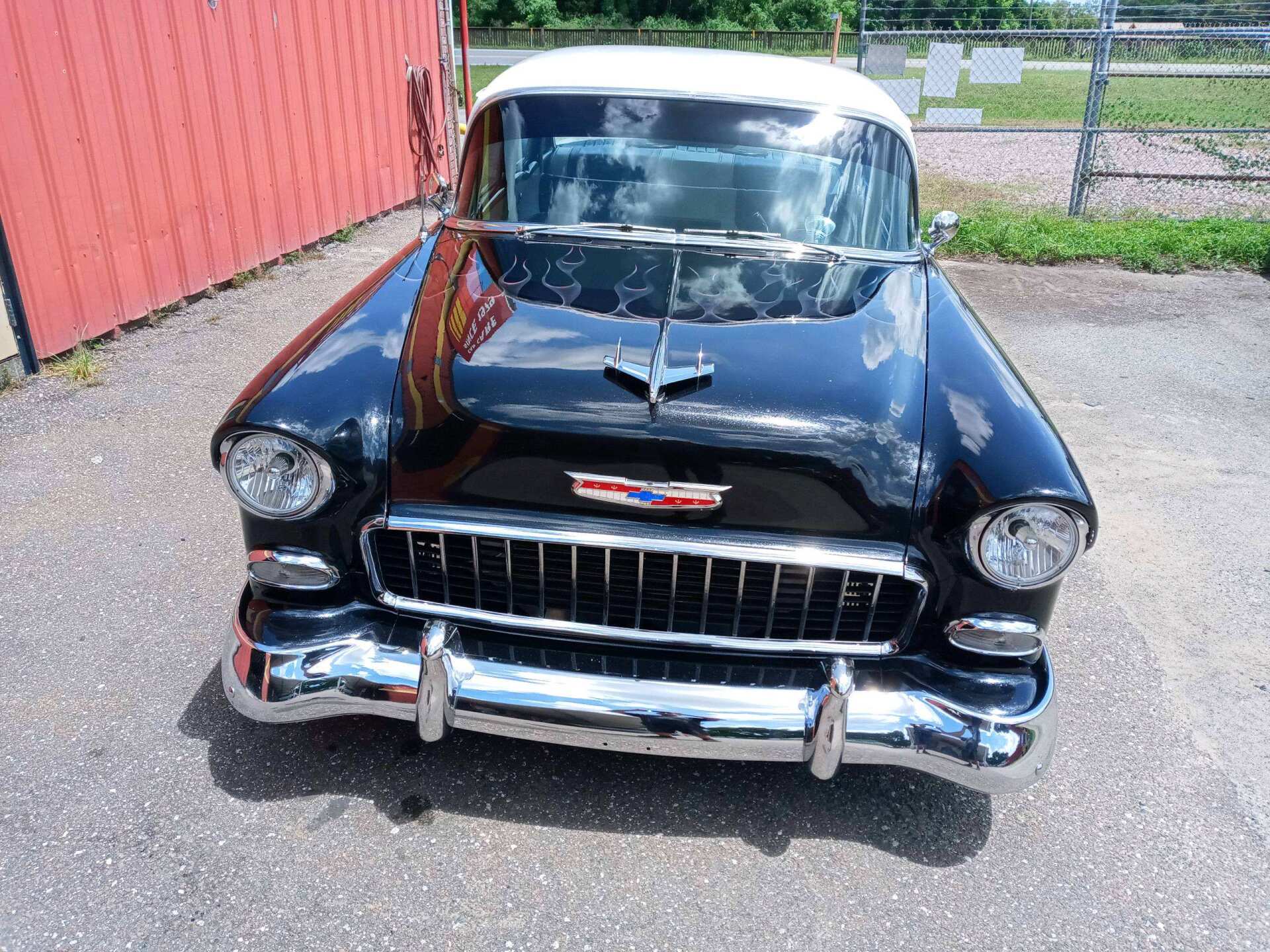 Front view of a shiny black classic Chevrolet parked on gravel beside a red building