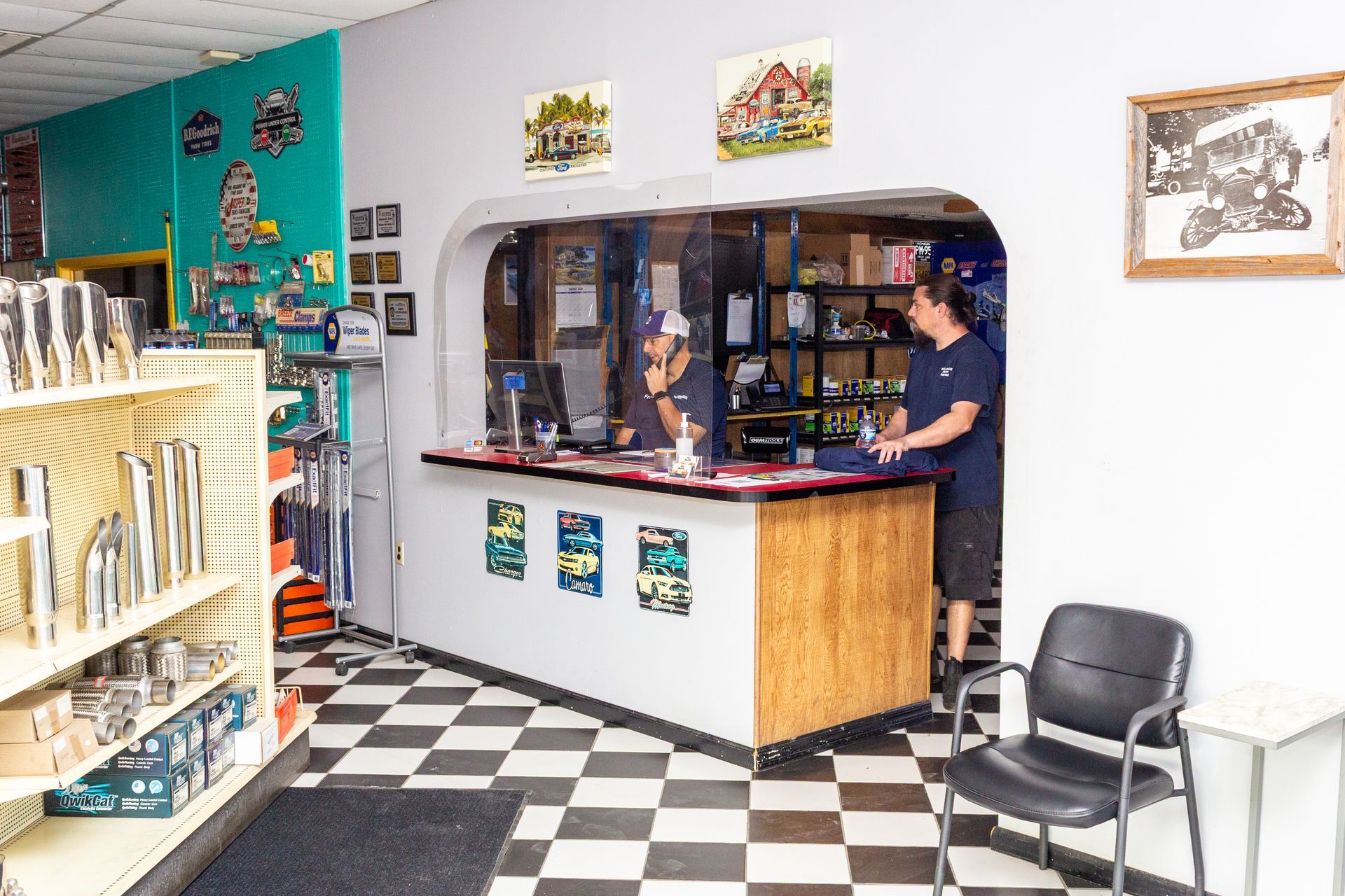 Small shop counter with a person standing behind it, black-and-white checkered floor, colorful shelves, and framed art.