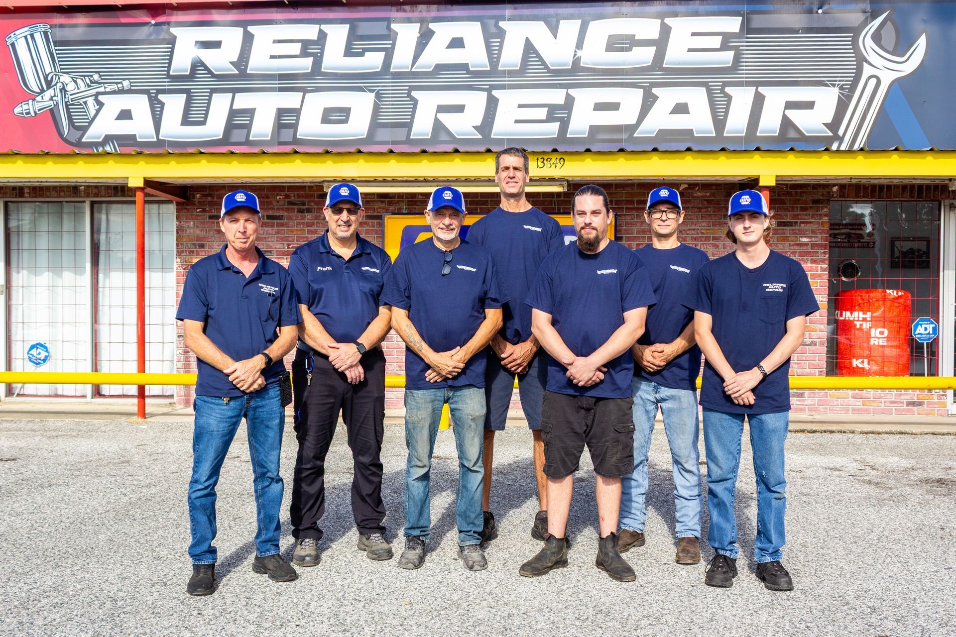 Reliance Auto Repair staff posing in front of the shop sign, wearing navy uniforms and caps.