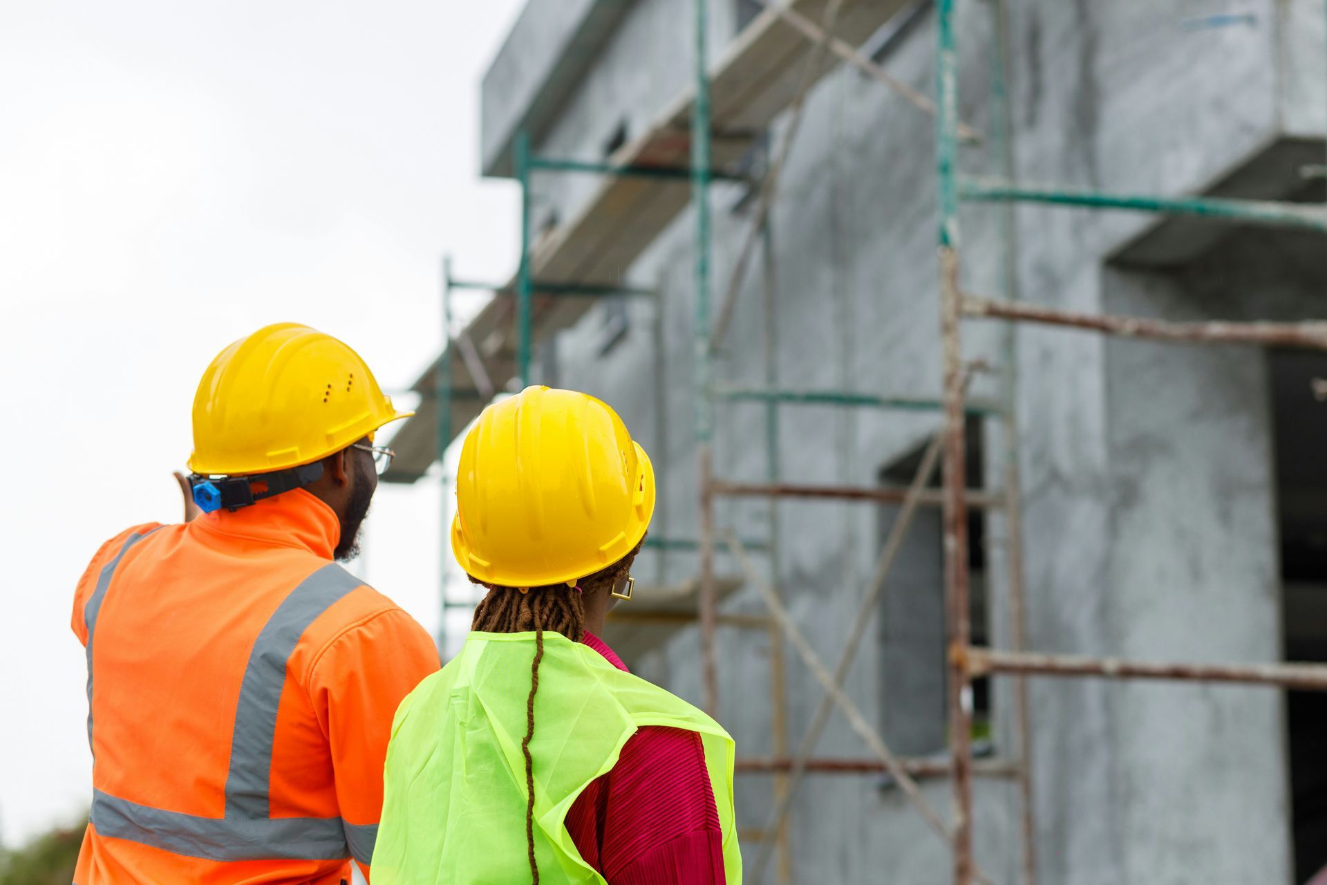 Two construction workers in hard hats and safety vests standing by a scaffolded building under construction