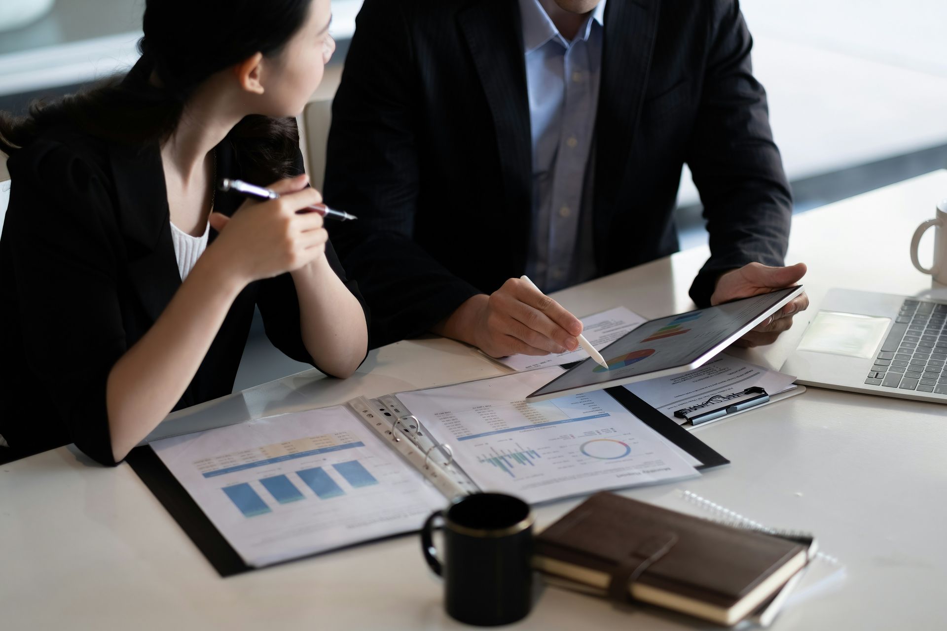 Two businesspeople reviewing charts and documents at a desk with a laptop and coffee mug