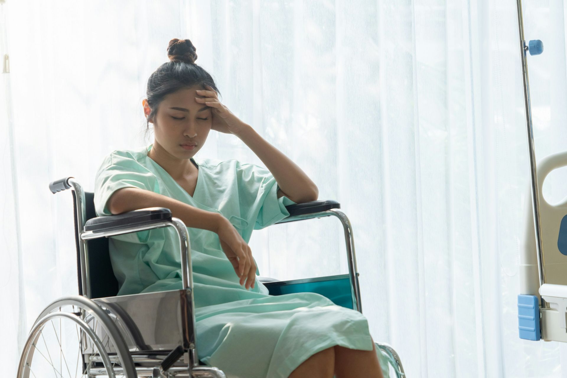 Woman in a wheelchair, holding her forehead and looking pensive in a bright hospital room.