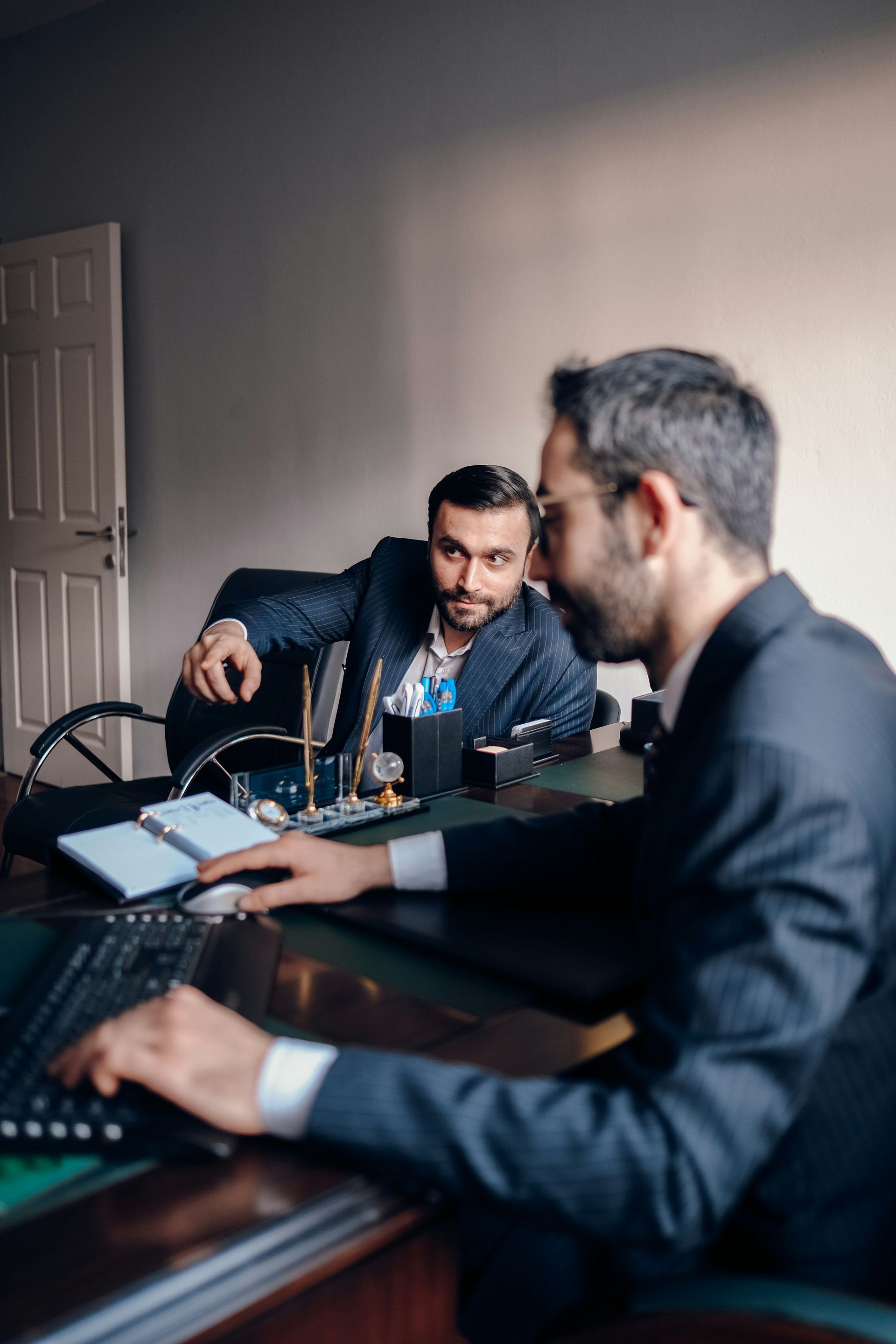 Two men in an office meeting, one typing on a laptop while the other speaks across the table.