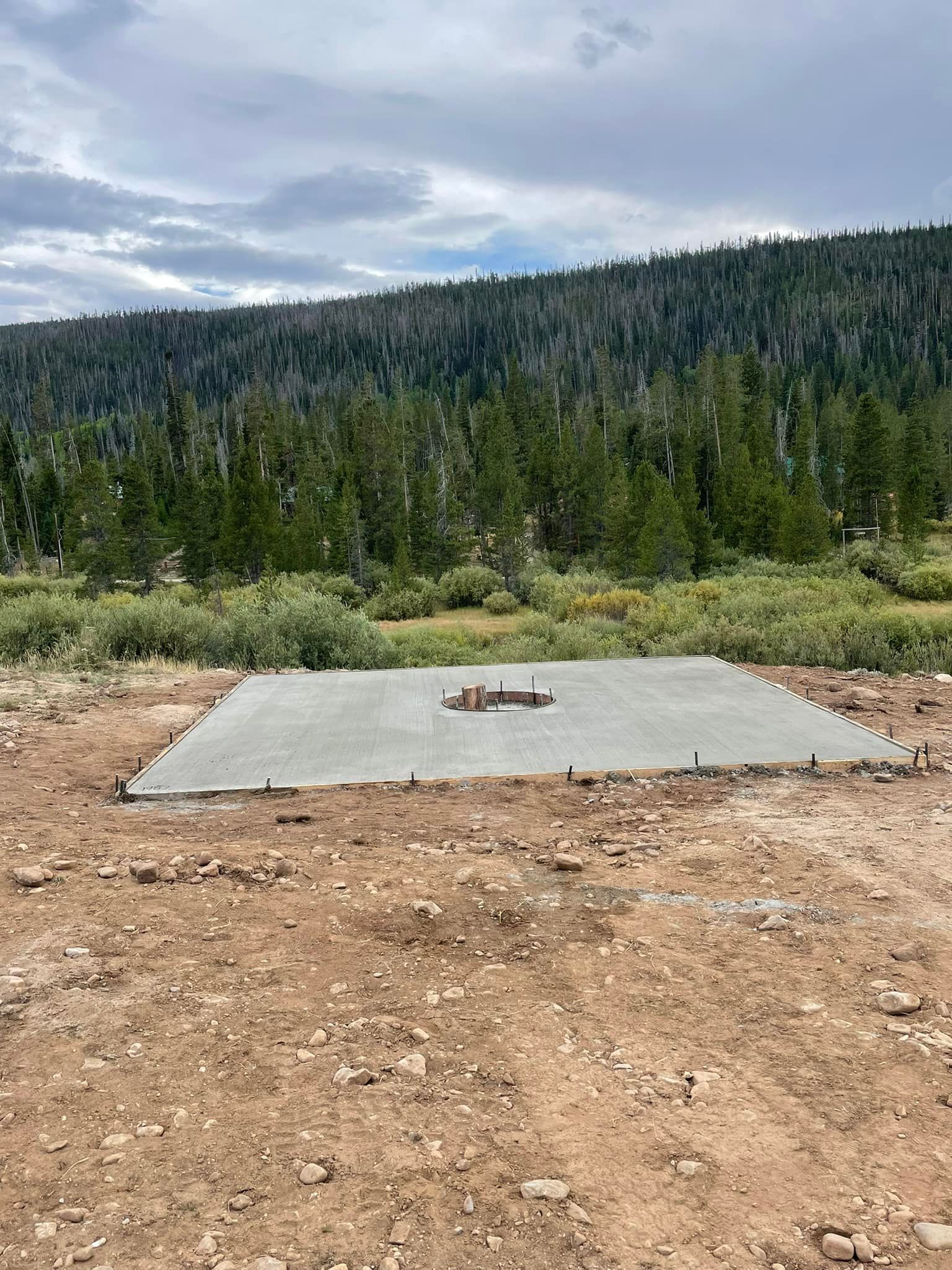 Newly poured concrete foundation in a natural setting, with vegetation and trees visible in the background.