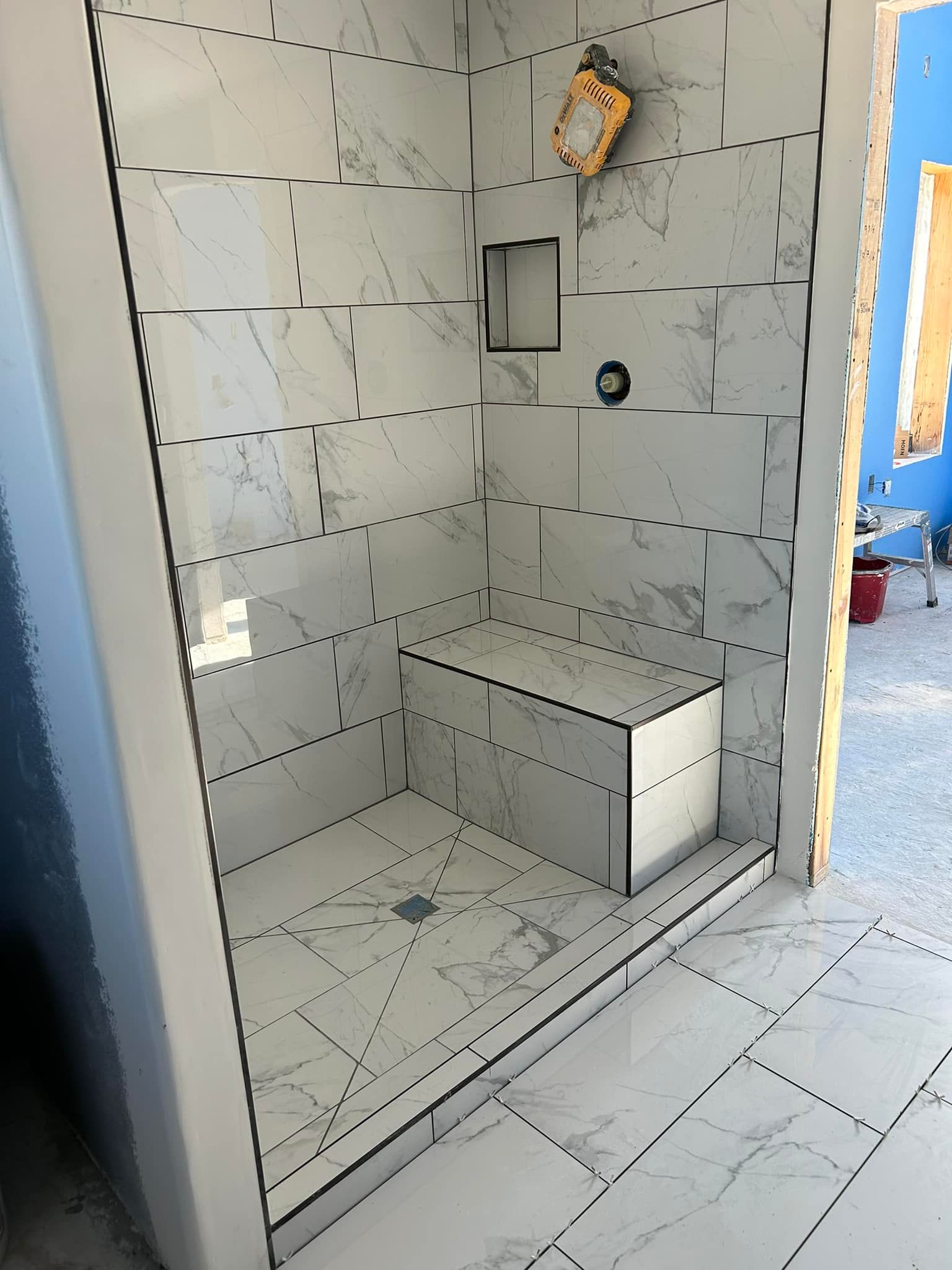 Finished white tile shower with a built-in seat and niche. Black grout lines and a drain.