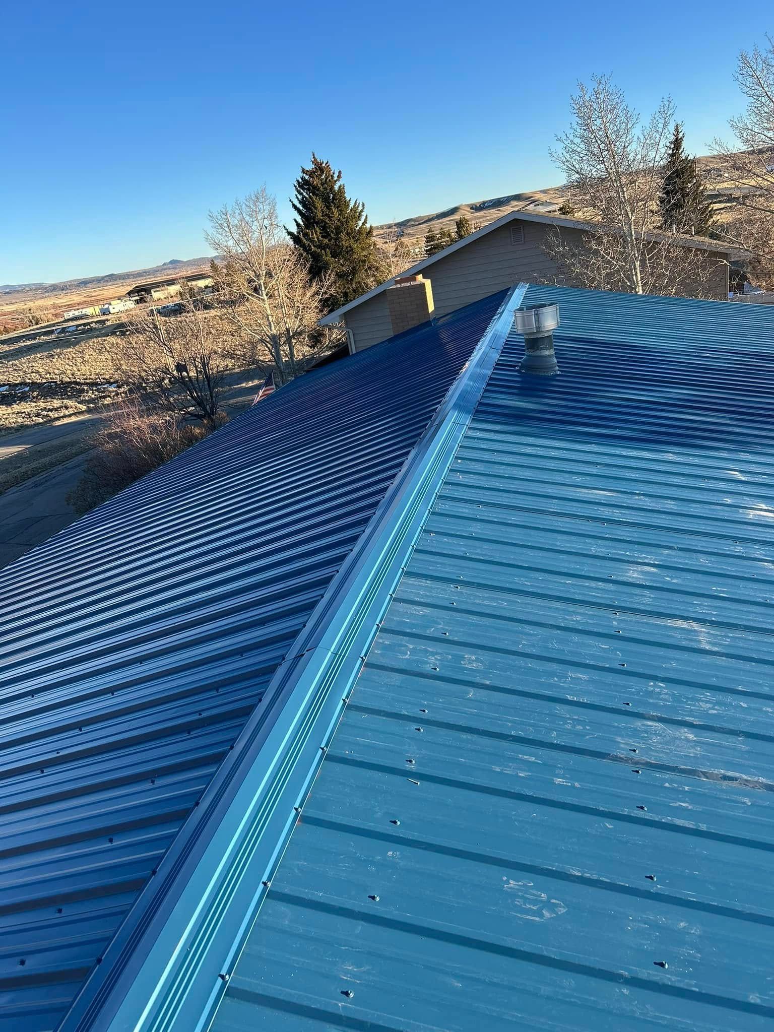 Blue metal roof with a ridge, set against a clear blue sky, overlooking a landscape with trees and hills.