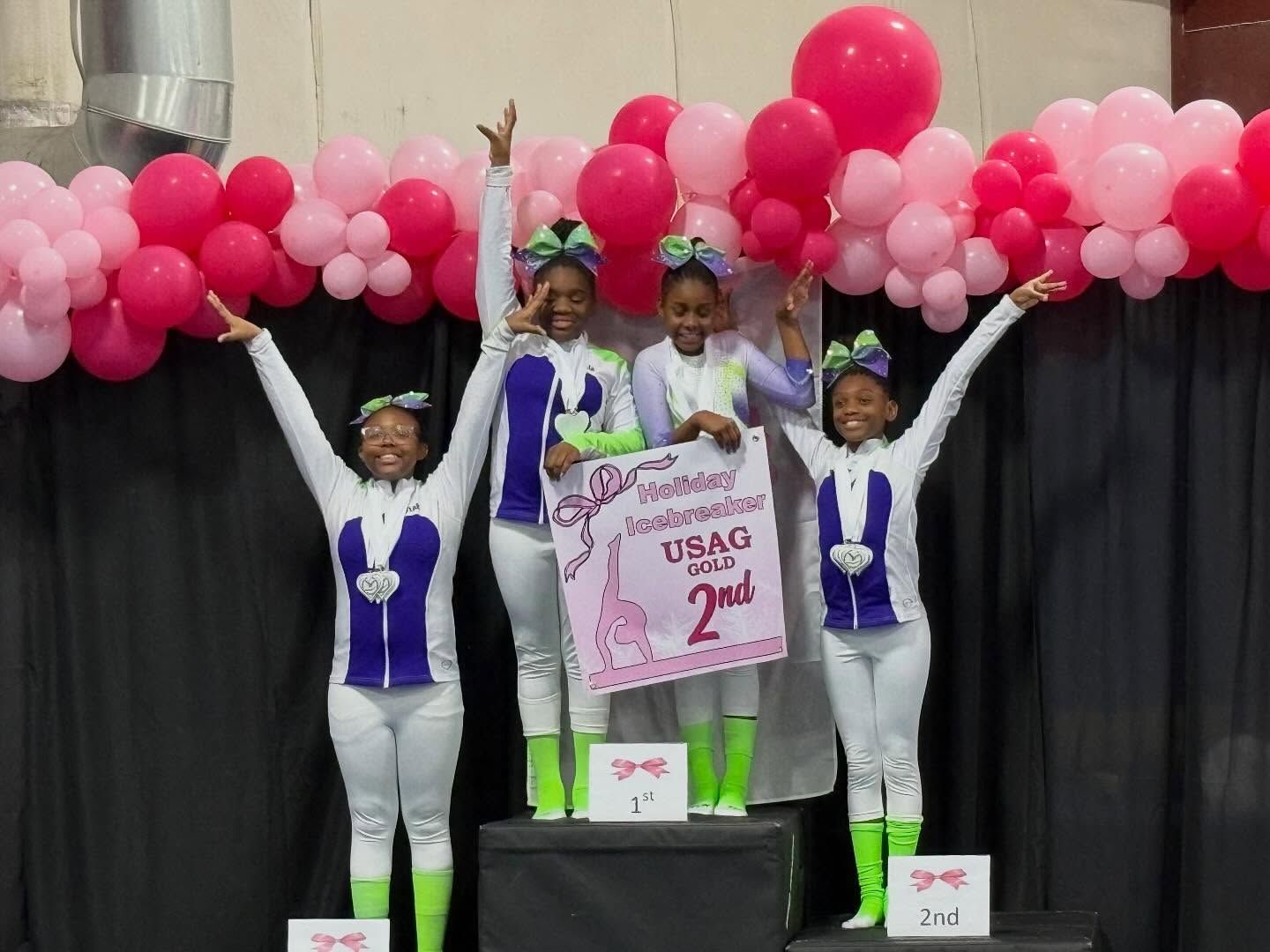 Four medalists on a podium with balloons, raising arms in celebration at a gymnastics award ceremony