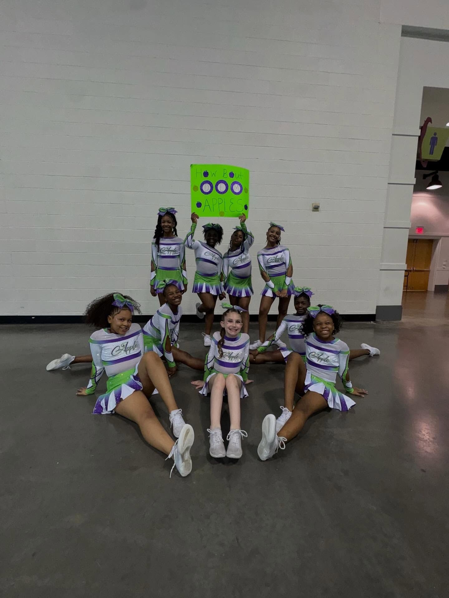 Cheer team in white and green uniforms posing on a gym floor with a bright green sign behind them