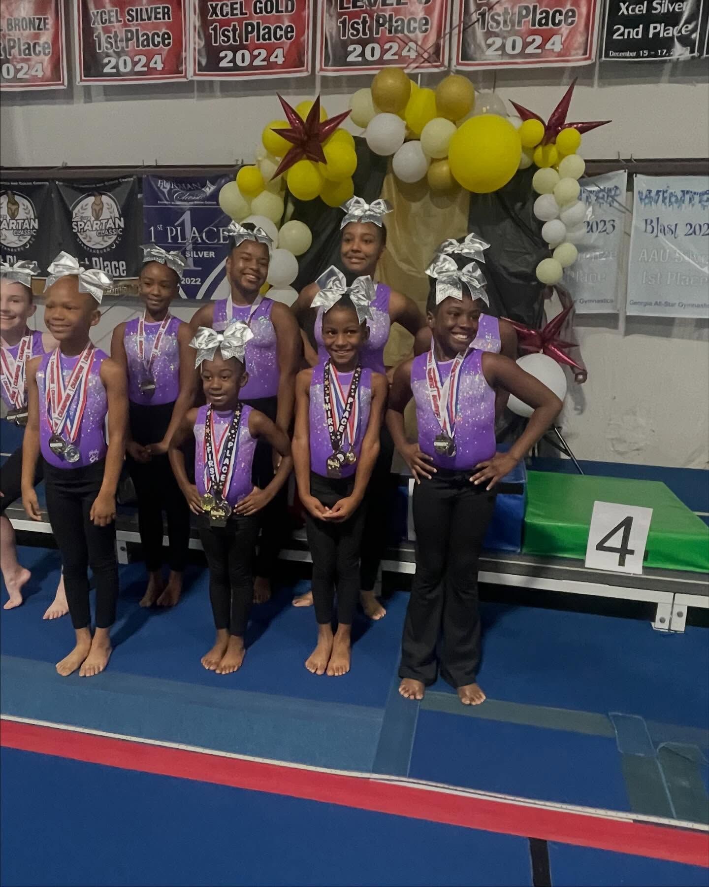 Young gymnasts in purple uniforms stand on a blue mat at a medal ceremony with balloon arch backdrop