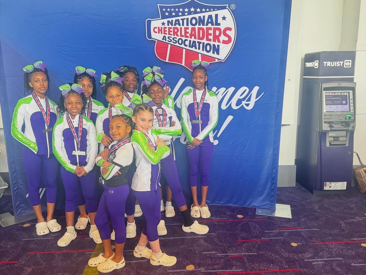 Cheerleading team in blue, white, and green uniforms posing in front of a National Cheerleaders Association banner.