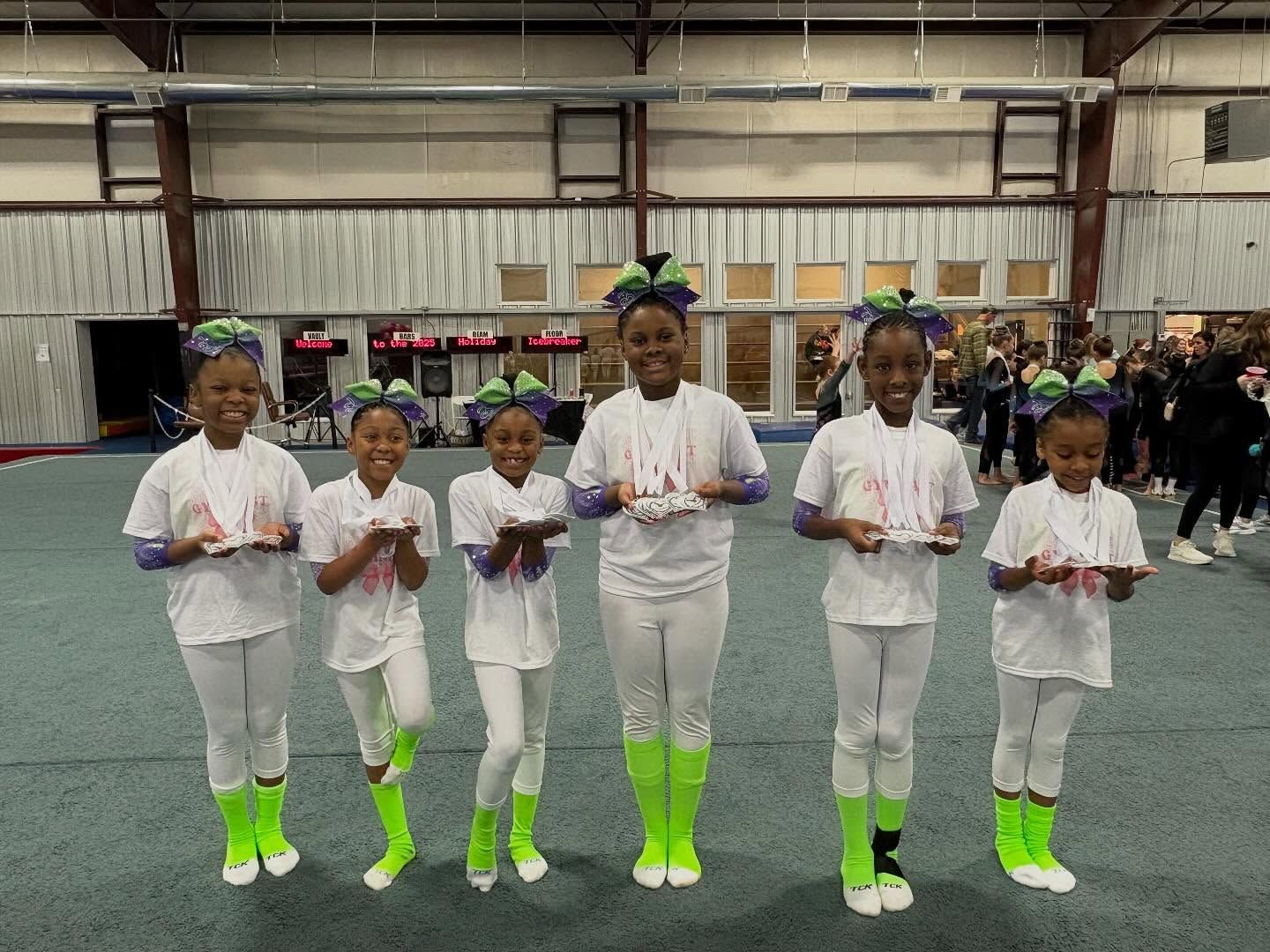 Six children in white and neon green outfits stand in a gym, holding cups and posing in a line.