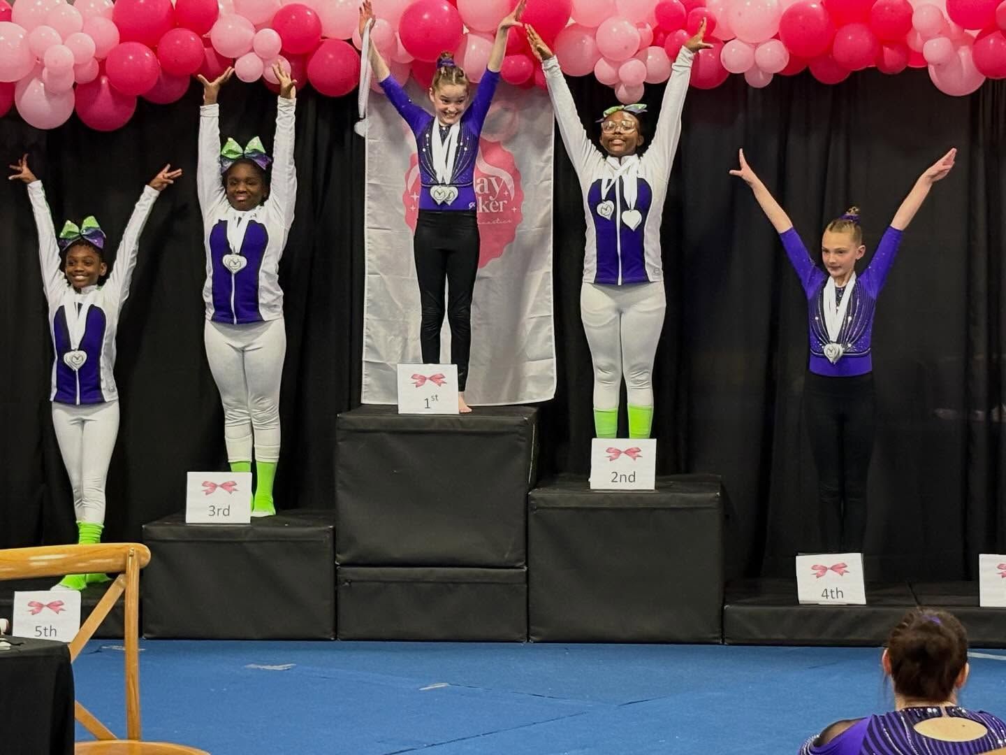 Gymnastics podium celebration with five athletes raising their arms under pink balloon decorations