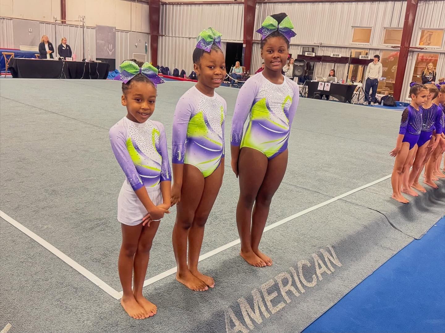 Three young gymnasts in purple and white leotards stand on a mat at a competition.