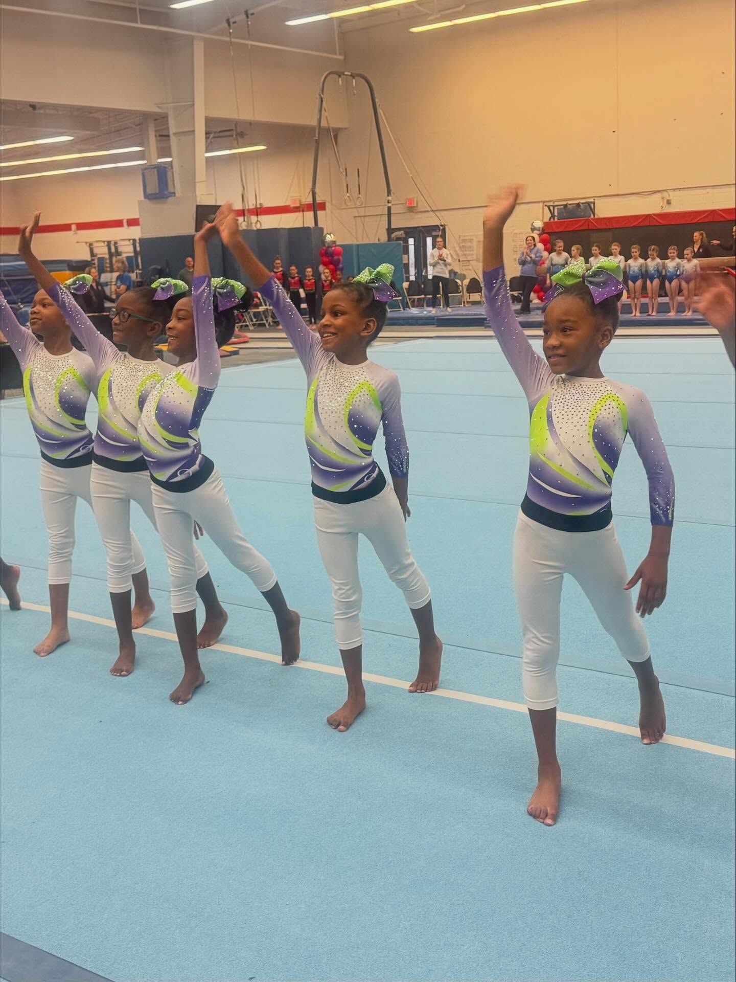 Four young gymnasts in matching leotards stand on a blue mat, arms raised in a gym.