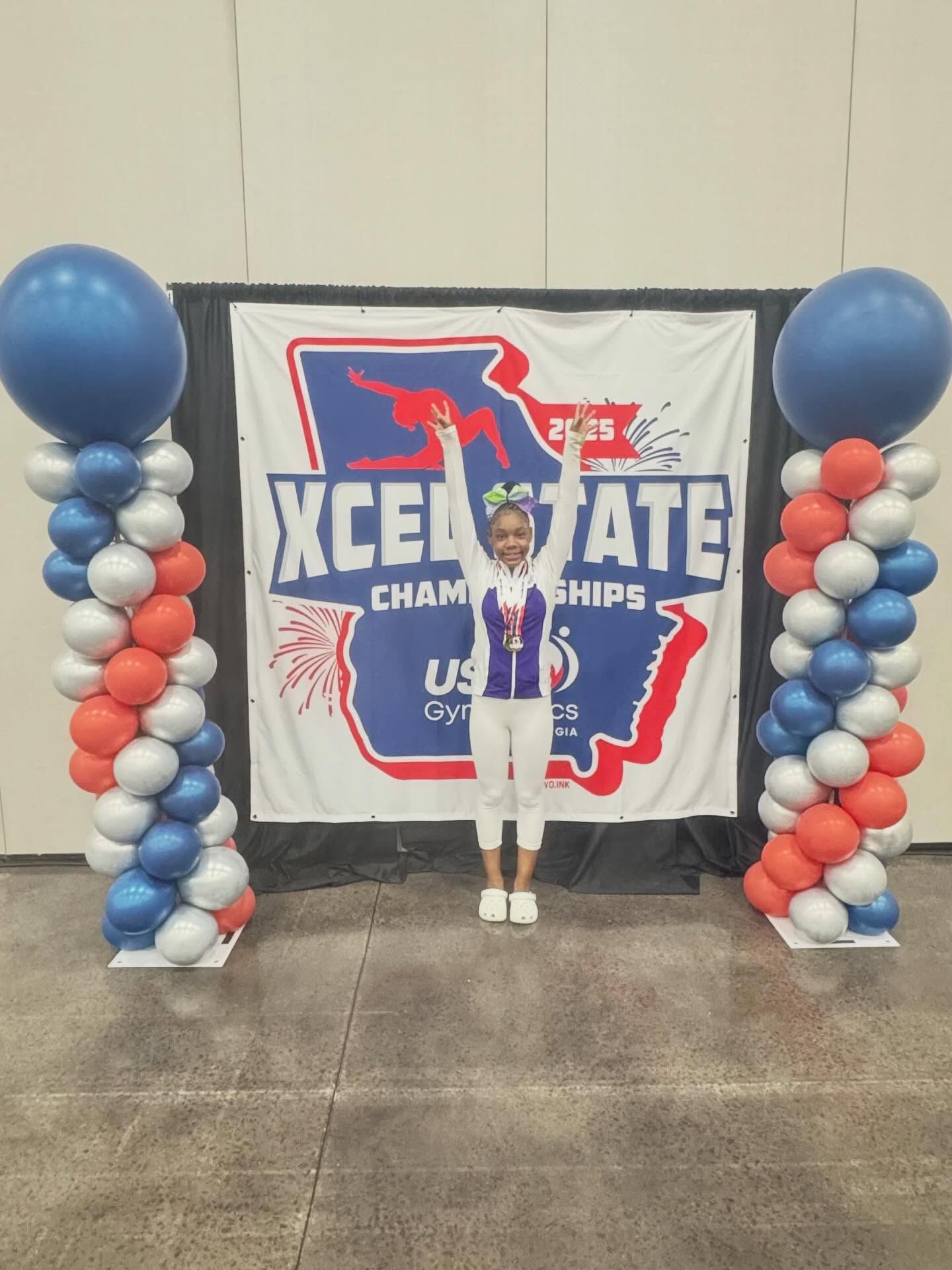 Young athlete posing with trophy arms raised between blue, white, and red balloon columns at a championship backdrop.