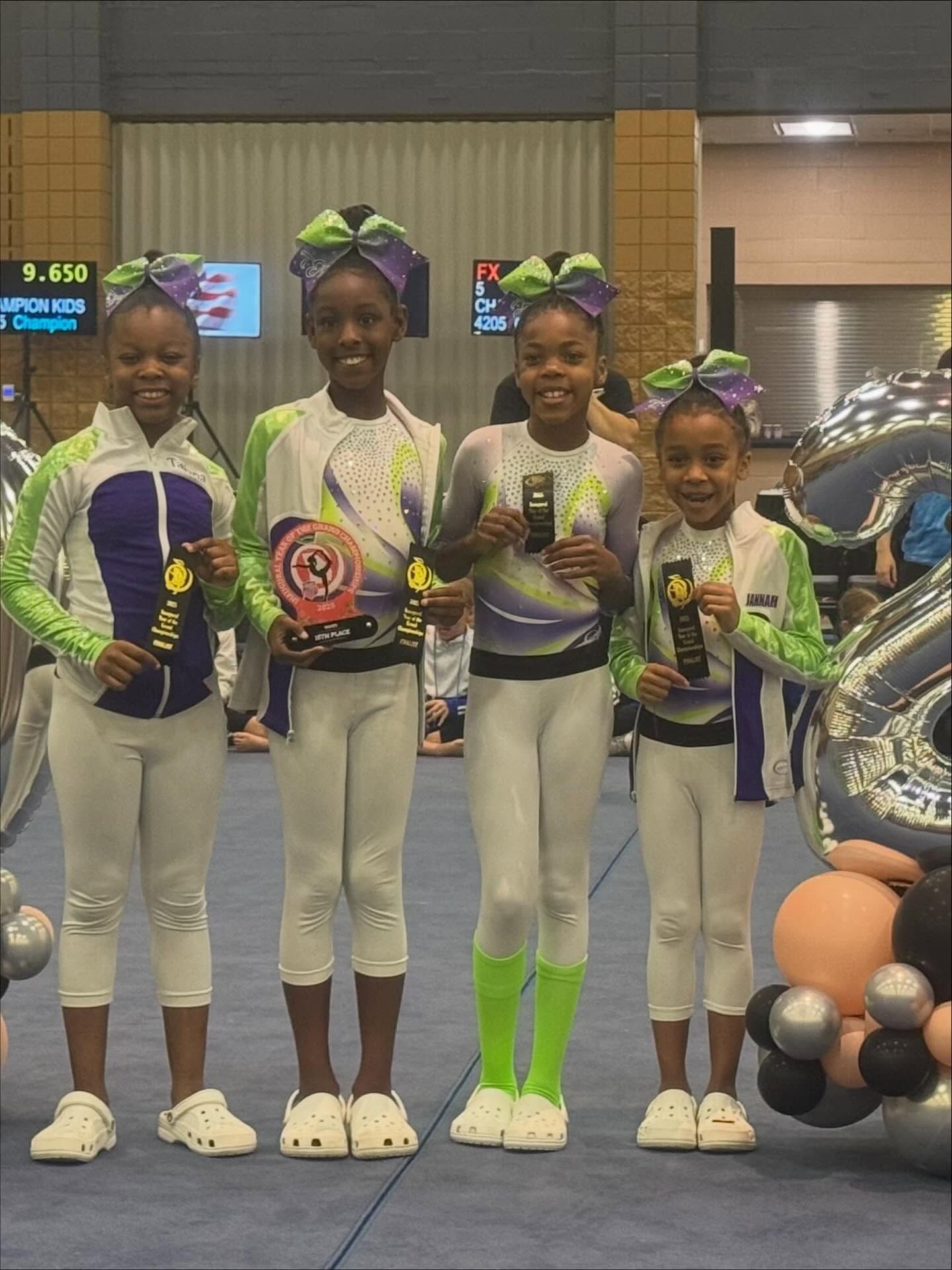 Four young gymnasts in white and neon uniforms pose with medals on a competition podium.