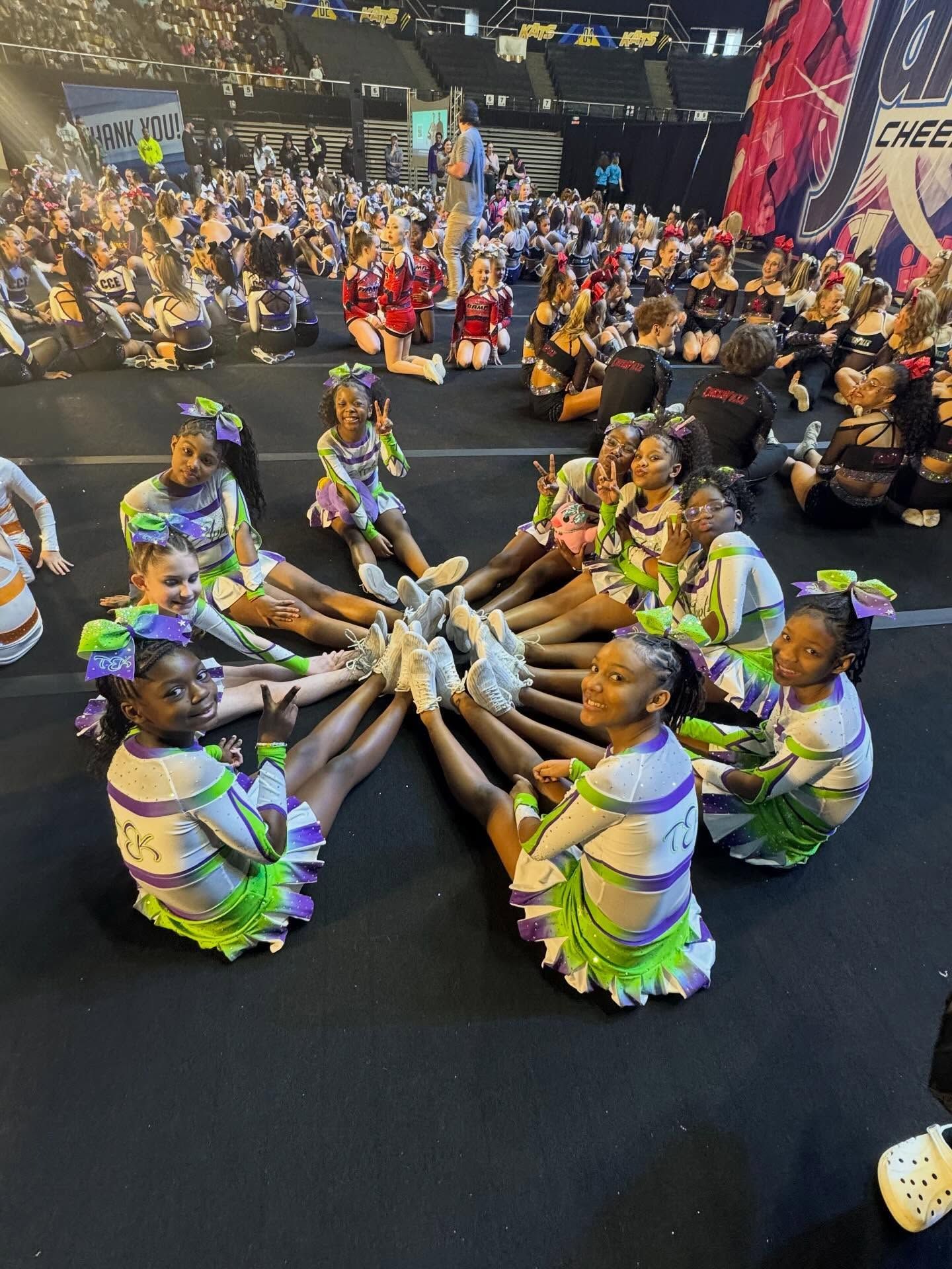 Dancers in colorful costumes sit in a circle, reaching feet together during a stage performance.