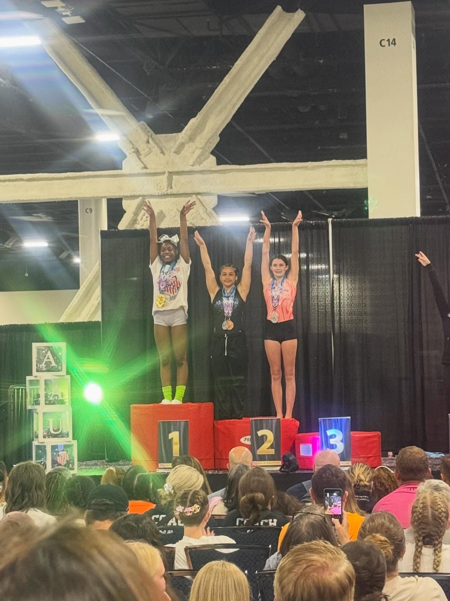 Three medalists on a podium raising their arms at an indoor event, with a cheering crowd in front.