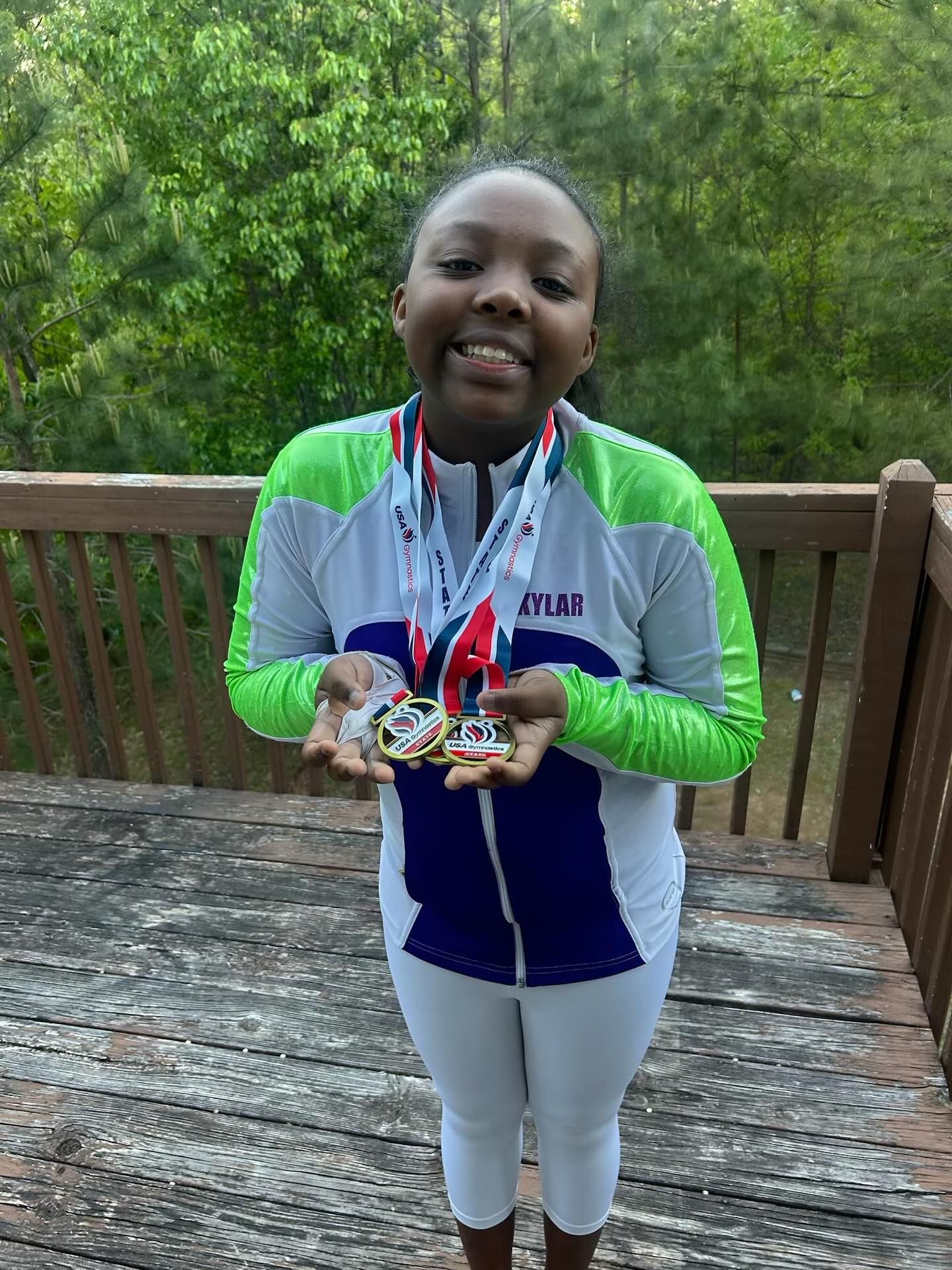 Child smiling and holding medals on a wooden deck with trees in the background